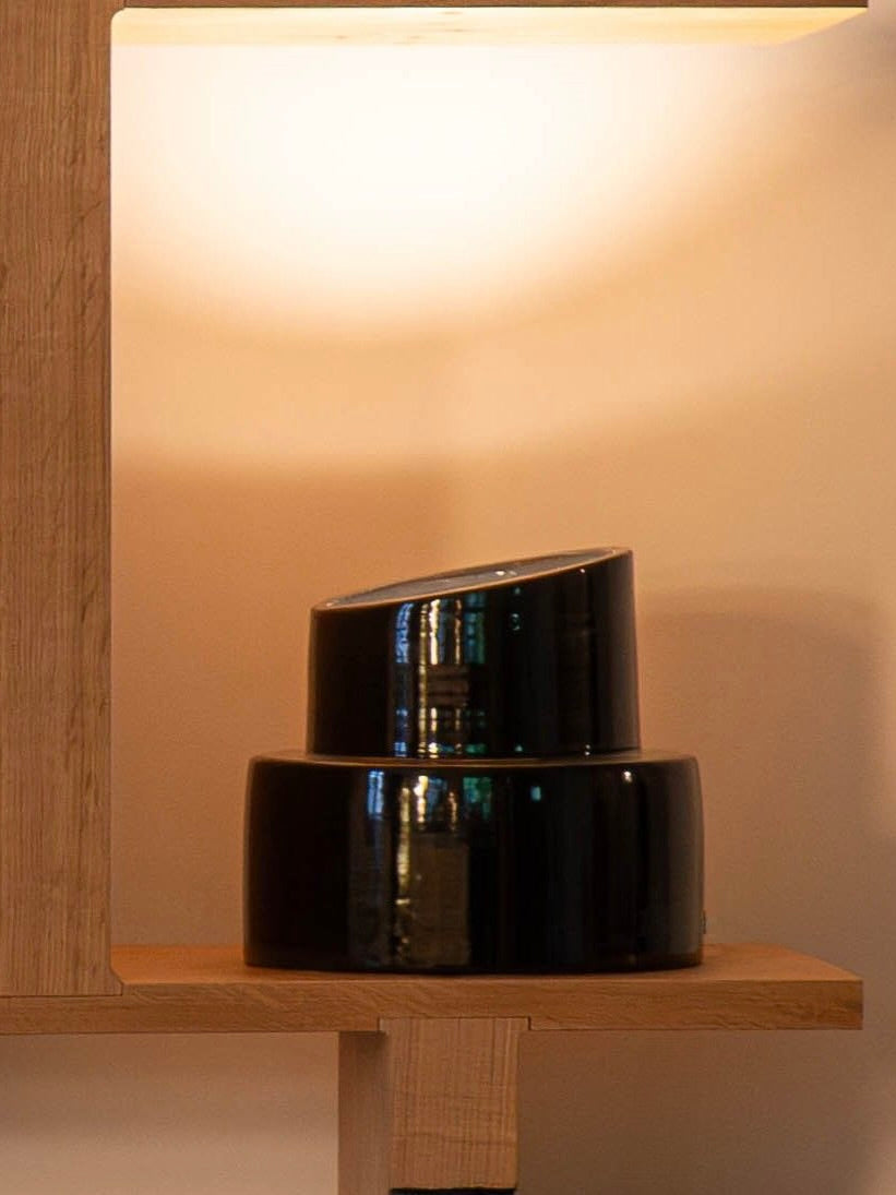 Two glossy black ceramic bowls of different sizes are stacked on a wooden shelf under the glow of a warm light from the COSEINCORSO "Pillar" Table Lamp above, giving the light wood-finished shelf a cozy ambiance.