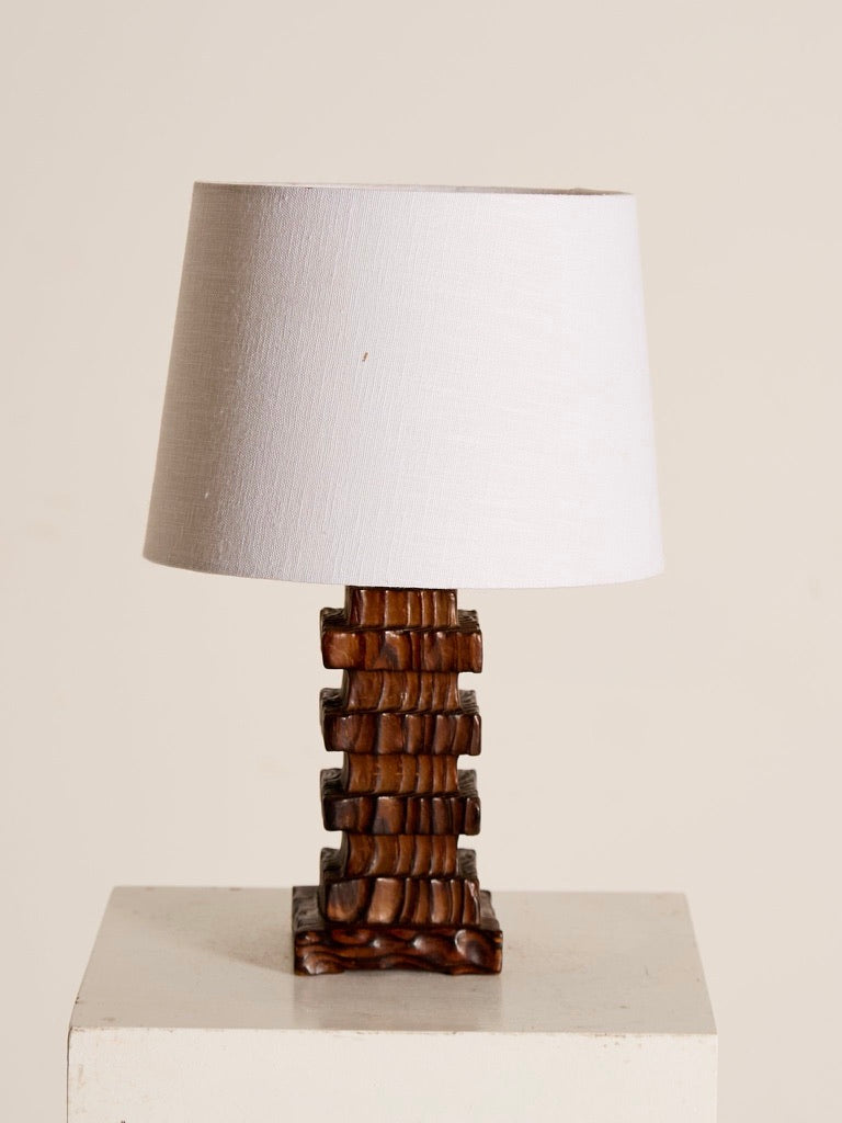 The Polish Pogda Lamp by Ancien et Jolie is a sculptural table lamp with a white shade and a base of stacked, carved wooden blocks in a geometric style reminiscent of 1960s Poland. Possibly made from combed Douglas fir, it rests elegantly on a white square platform against a plain backdrop.