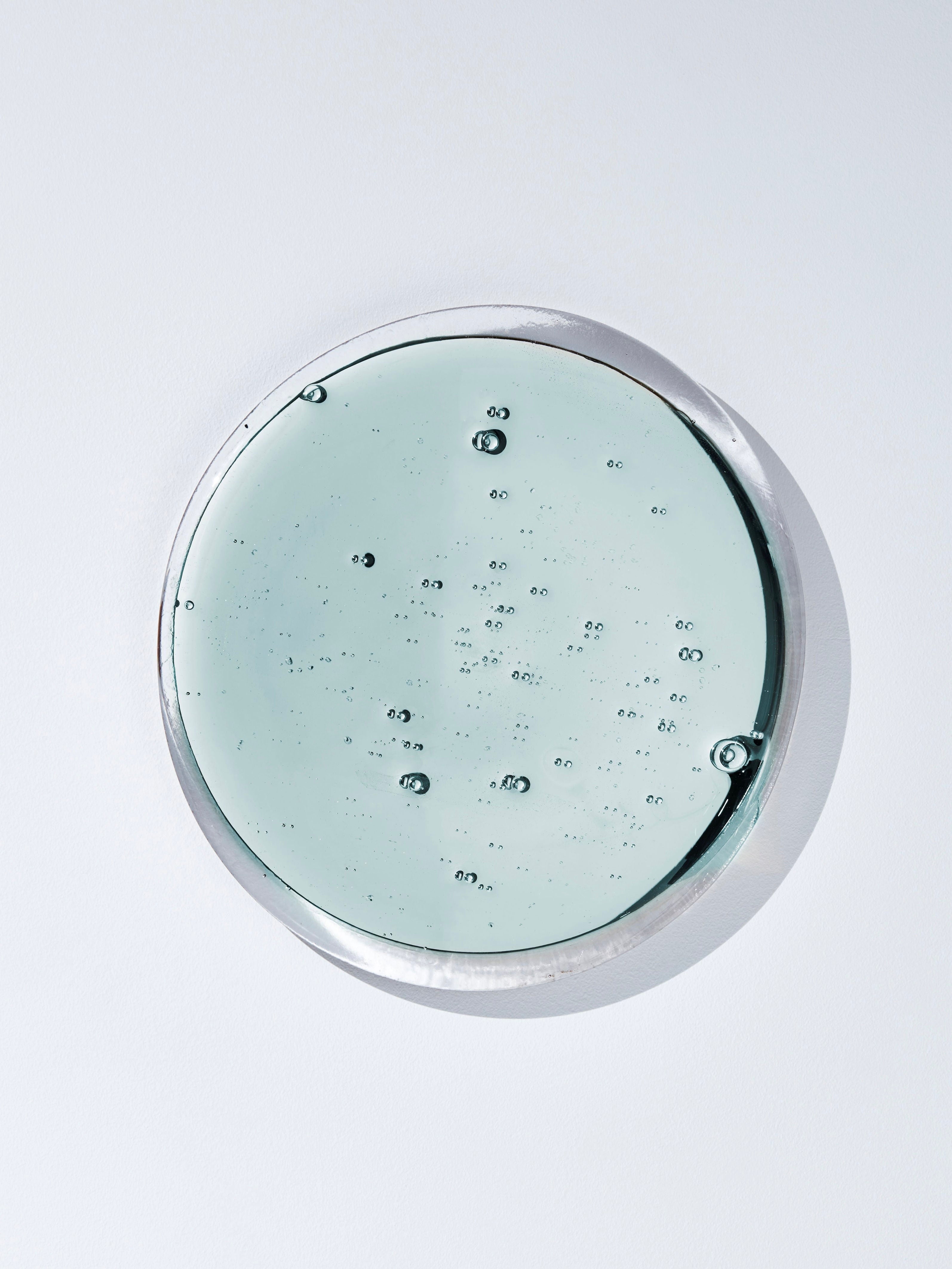 A close-up view of TĂȘte Studio's Puddle Mirror, displaying a clear, gel-like substance with tiny bubbles scattered throughout, resembling water droplets. The Puddle Mirror sits on a white surface, and its rim is slightly reflective like mirrorized glass.