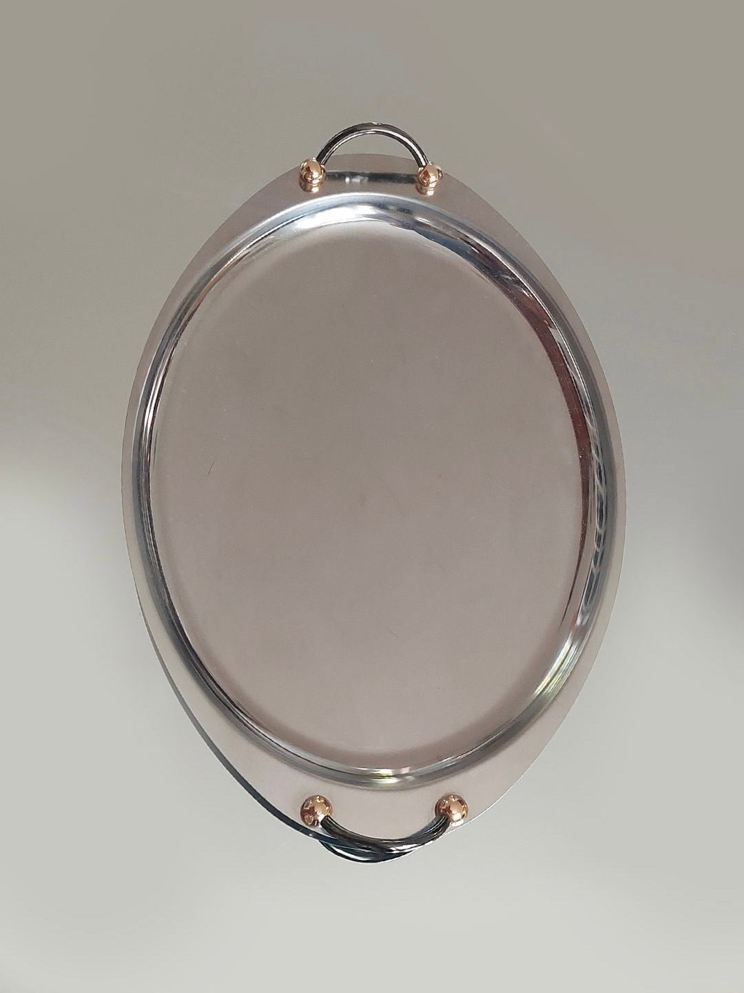 A Medium Oval Steel Tray by Les Objoies, featuring elegant gold handles and two black grips, is centered against a plain light grey background. The minimalist design boasts a smooth, glossy surface and a slightly raised edge.