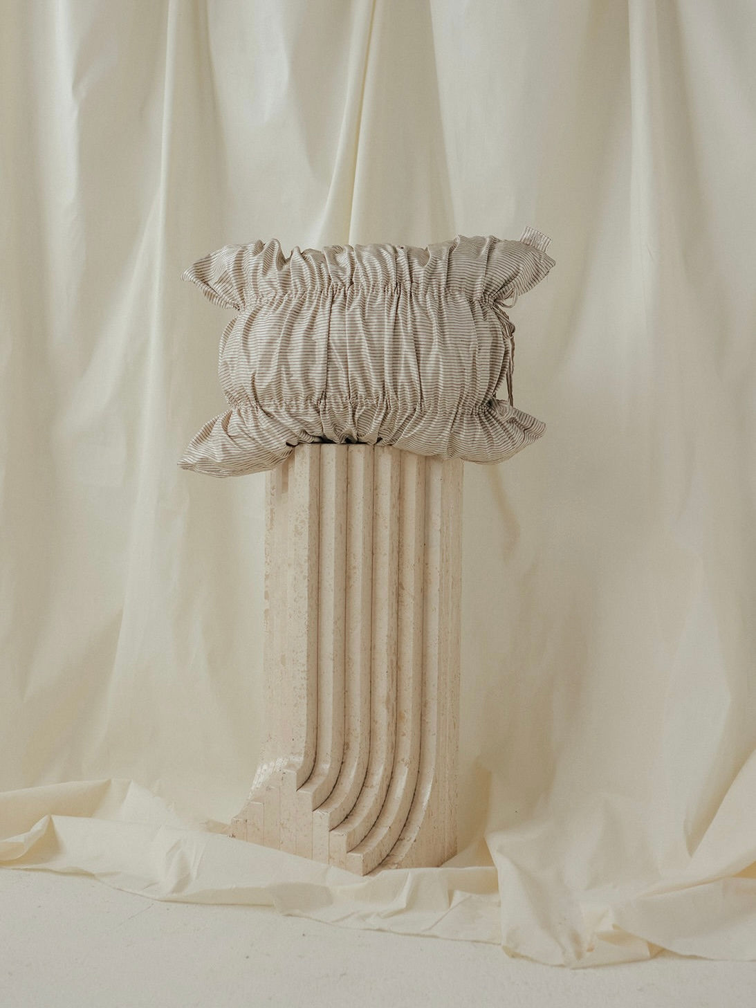 A beige Daphne Striped Cushion by SANTA LIVING, boasting a ruffled edge, rests on a sculpted marble pedestal against a softly draped cream fabric backdrop.