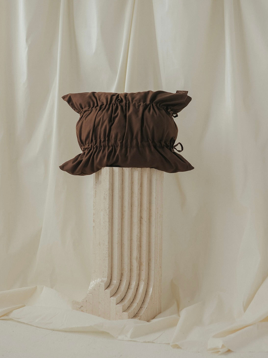 A SANTA LIVING Daphne Cocoa Cushion with drawstring edges is showcased on a textured white fluted column pedestal, set against draped cream fabric.