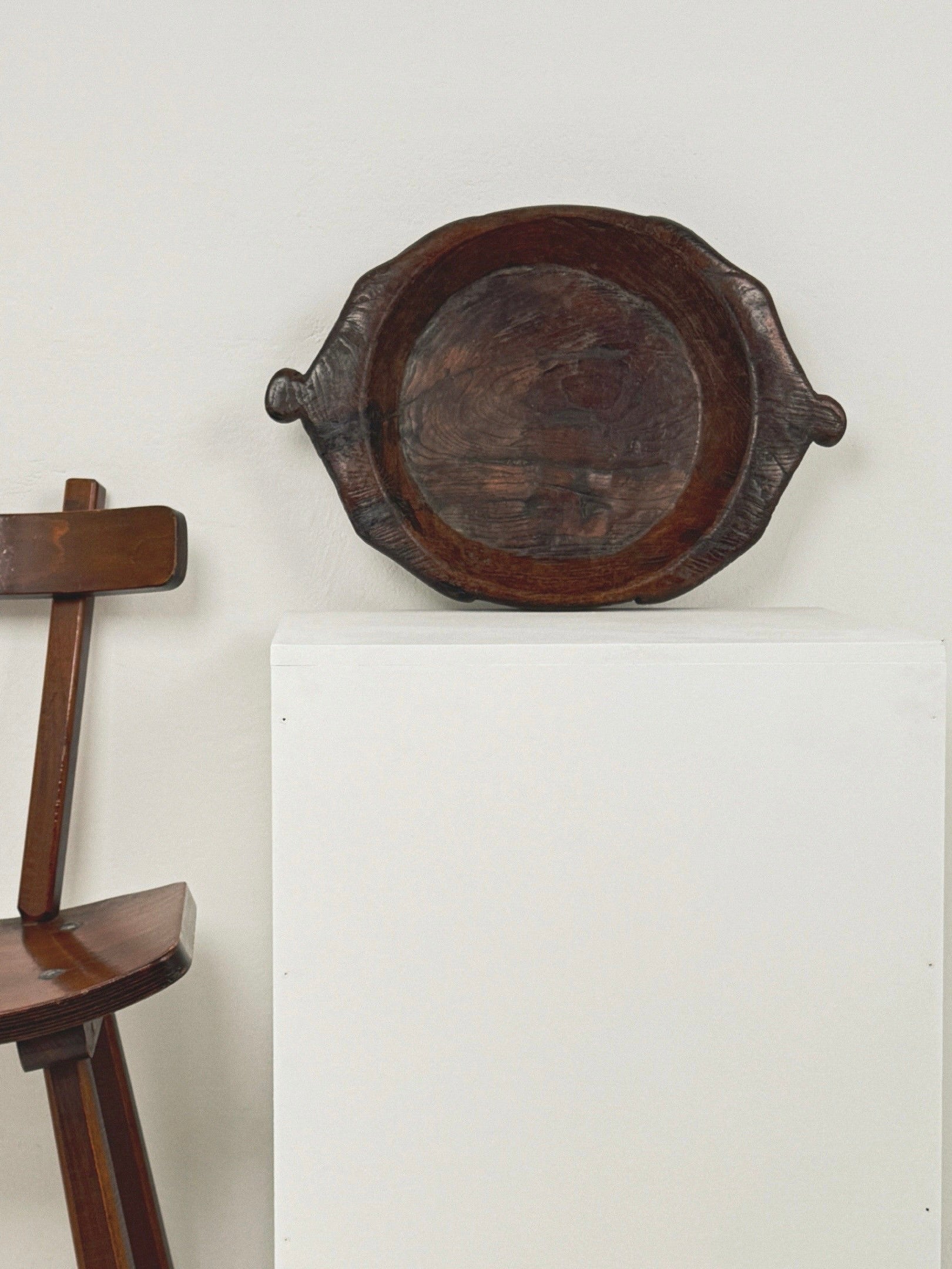 A Large Monoxyle Bowl from the 19th century by Figuresdesign, crafted from dark wood with handles, sits on a white pedestal, evoking French folk art. Nearby, a wooden chair's backrest is partially visible against a plain light-colored wall.