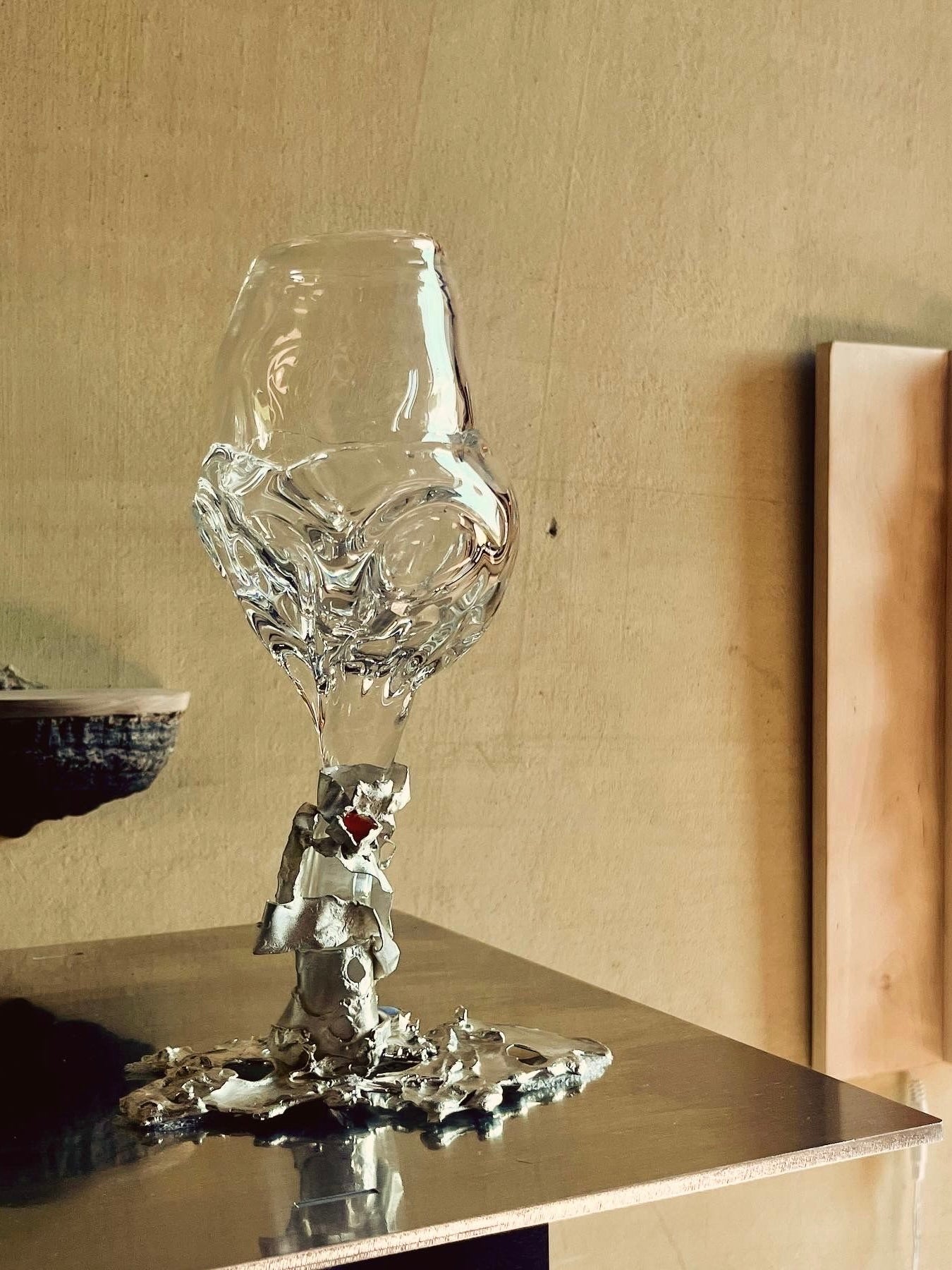 The Jewelled Goblet no. 9 by Lindsey Fontijn, crafted by Szkło Studio, is a transparent, hand-blown glass sculpture featuring a twisted veil design atop a metal base. Its fluid form gives the illusion of the glass melting seamlessly into the base. This exquisite piece is showcased on a metallic surface against a concrete wall with a wooden panel beside it.