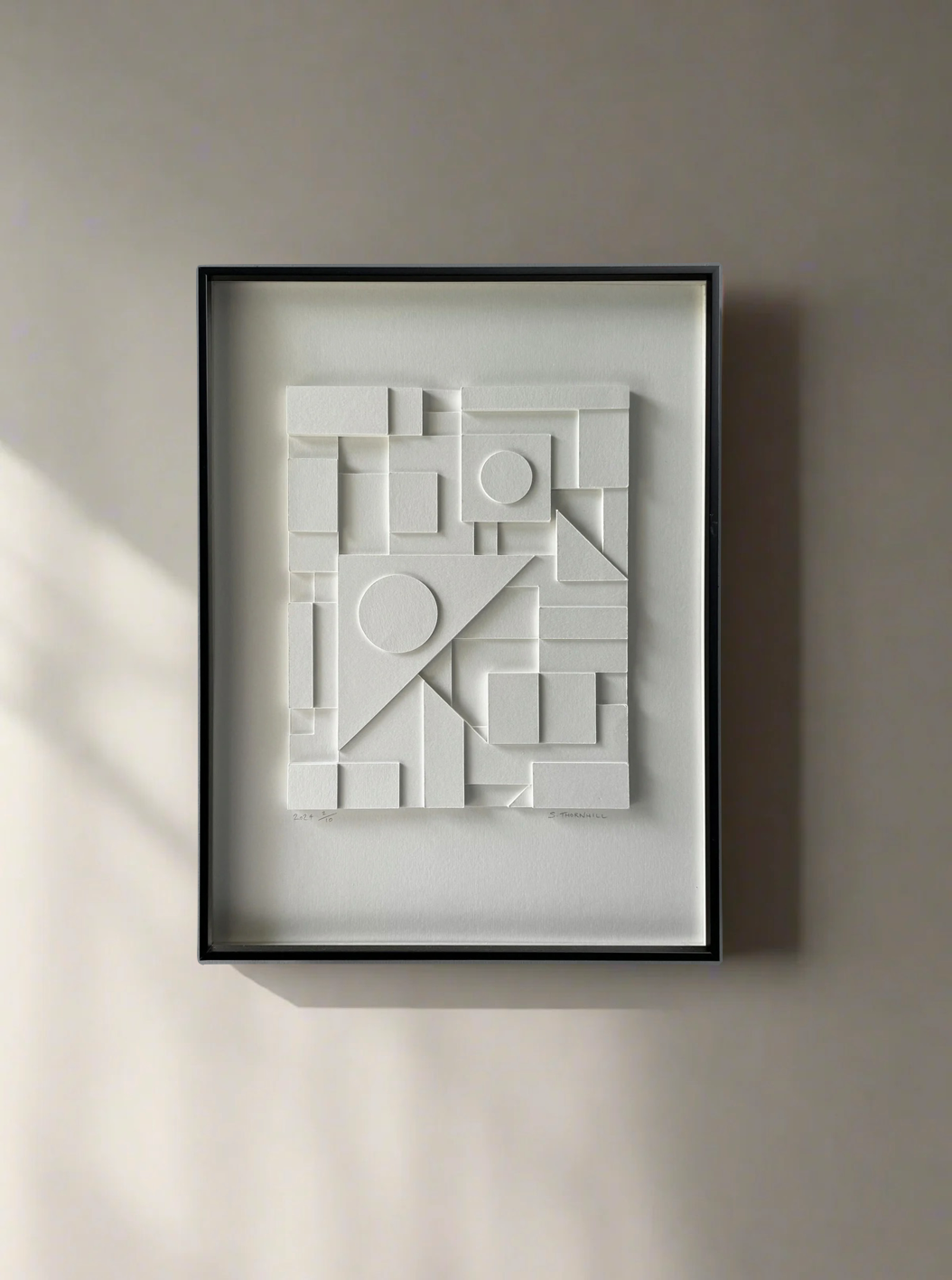 A minimalist artwork framed and hung on a wall. The piece, **Composition124** by **Sean Thornhill**, features geometric shapes in white—rectangles, circles, and triangles—creating a three-dimensional, abstract composition. The background is off-white, with soft, natural lighting highlighting its geometric style.
