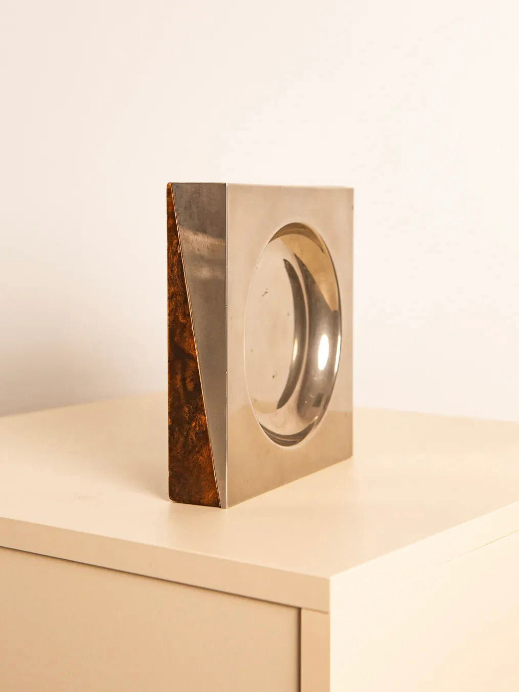 The Square Vide-poches 60's by Treaptyque is a geometric, silver-toned decorative piece with a circular cutout, featuring a reflective surface and a beautiful patina. It is elegantly showcased on a light-colored wooden table against a simple background, capturing the essence of Italian tradition.