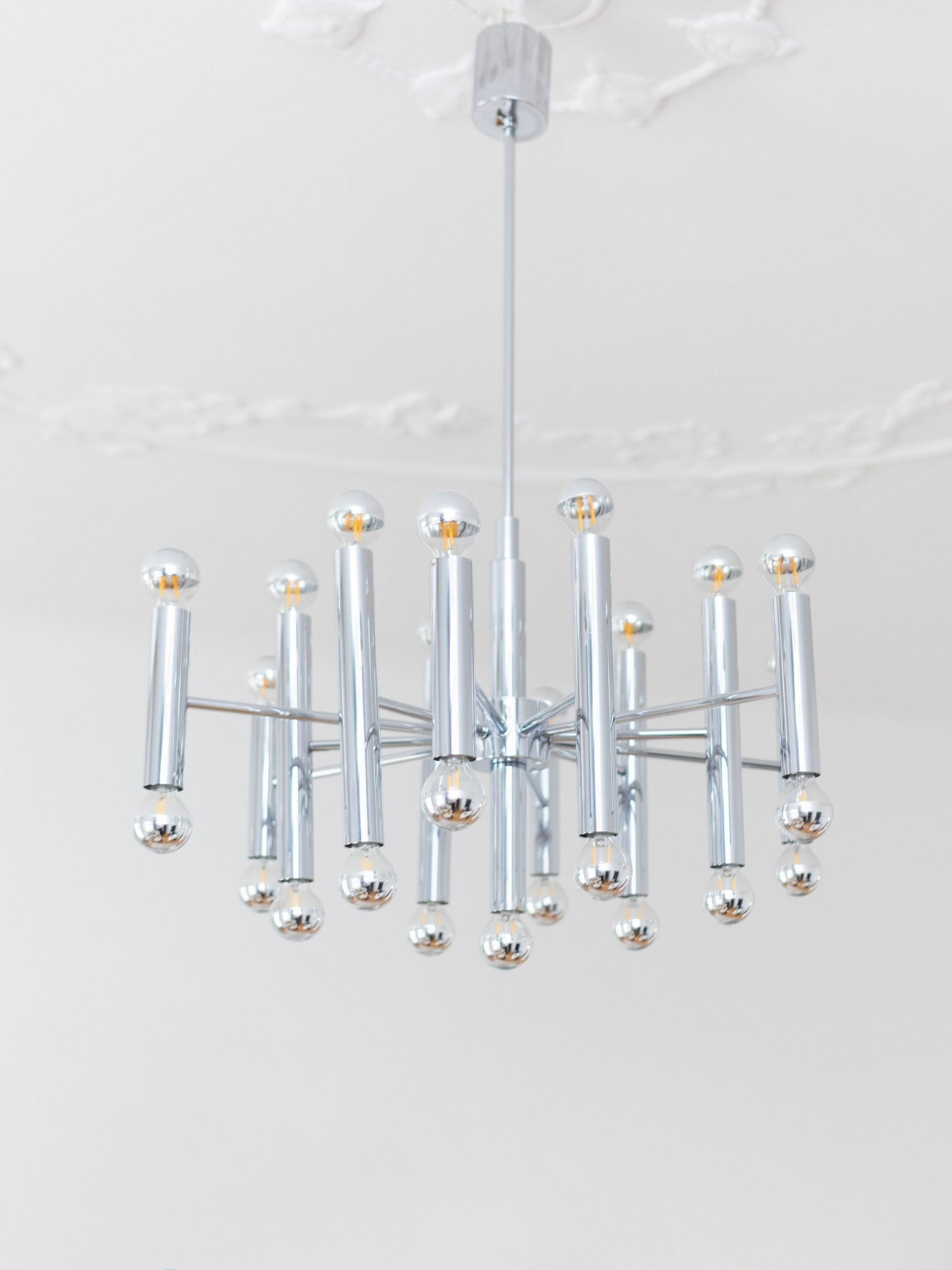 A Bottega Jacobs Gaetano Sciolari for Boulanger Chrome Ceiling Lamp, featuring its iconic cylindrical arms and spherical bulbs, elegantly hangs from a white ceiling adorned with ornate detailing, showcasing traditional craftsmanship.