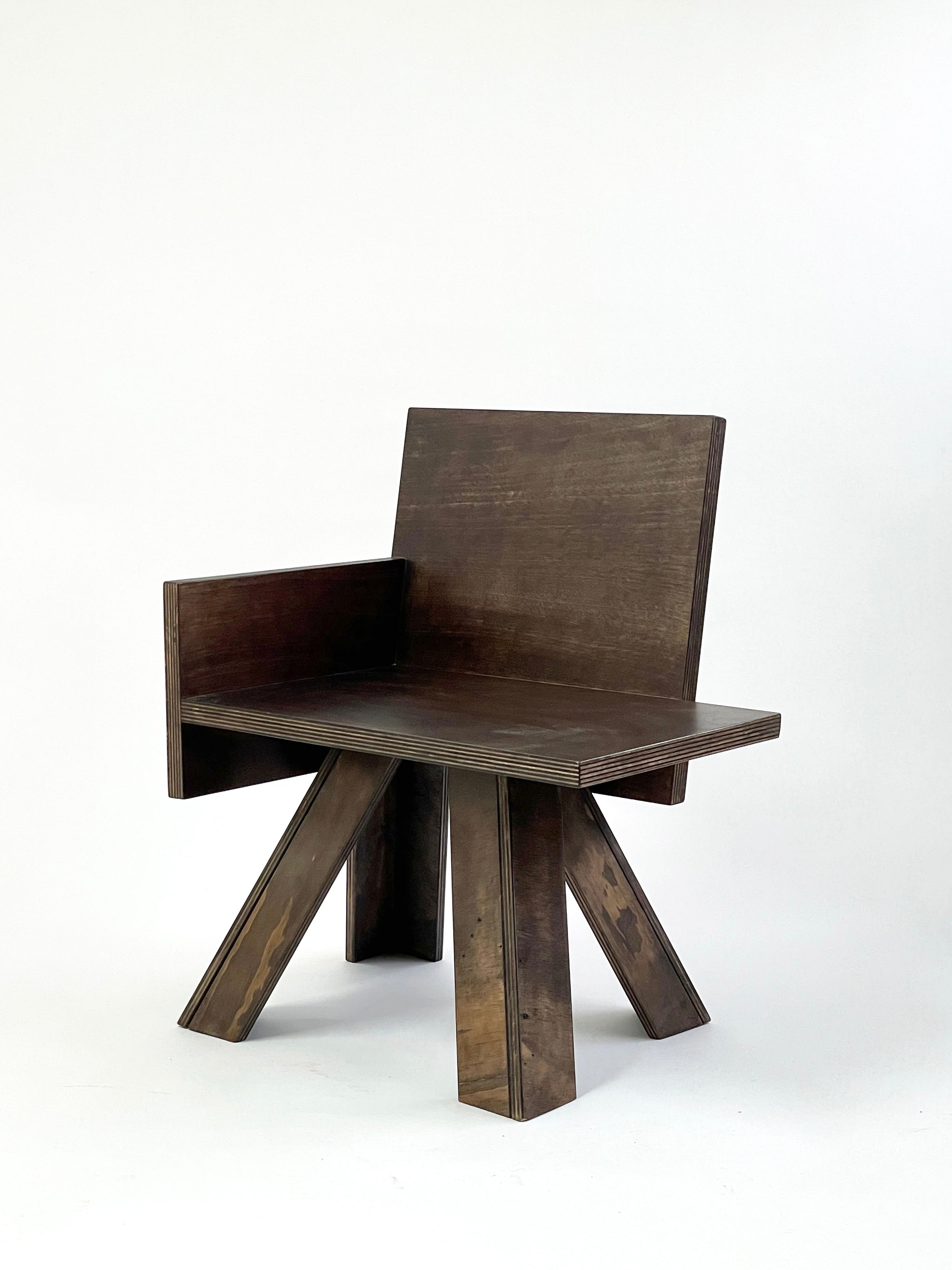 Presenting the End Table Chair by GOONS: a contemporary wooden chair that embodies geometric design and showcases expert craftsmanship. It features a rectangular backrest and seat supported by four angled, triangular legs against a simple white backdrop. Crafted by hand from reclaimed wood, its dark finish beautifully accentuates the intricate texture.
