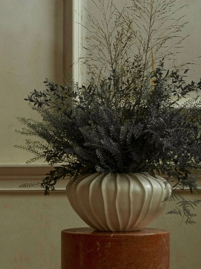 A white, ridged vase from Ransom & Dunn's Wide Cloak Vase collection is elegantly displayed on a pedestal, holding an arrangement of leafy branches and grasses in muted green and brown tones against a beige wall. This stunning piece is proudly handmade in Portugal.