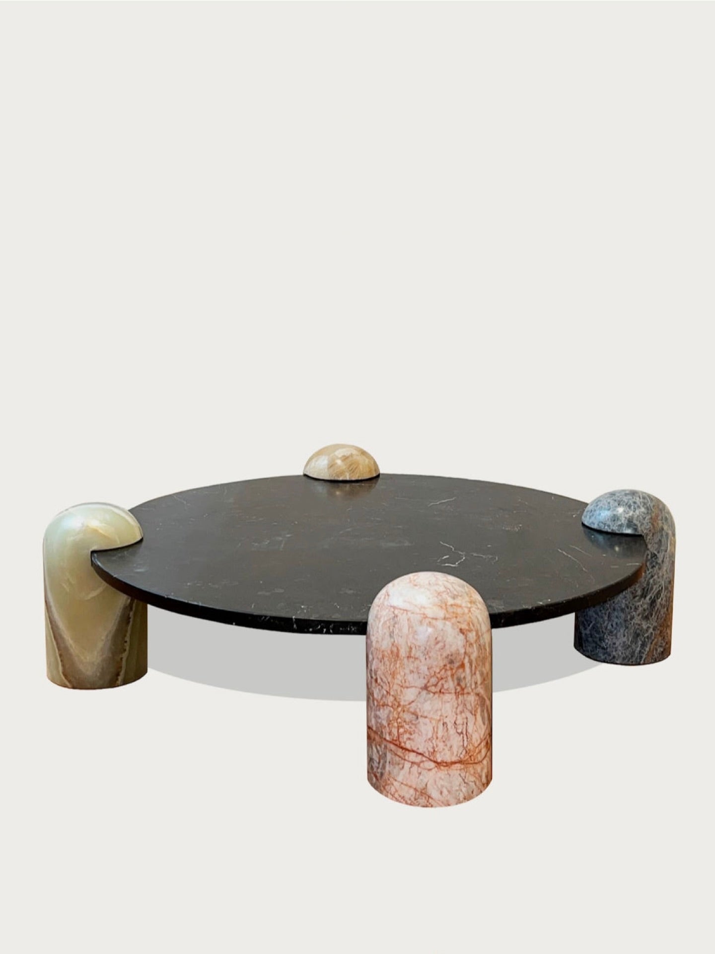 The Playtime Toy Table by Panorammma features a round black tabletop with sleek design and four rounded, pill-shaped legs in pink marble, beige, blue calcite, and light brown. A small dome made of similar marble sits atop the surface.