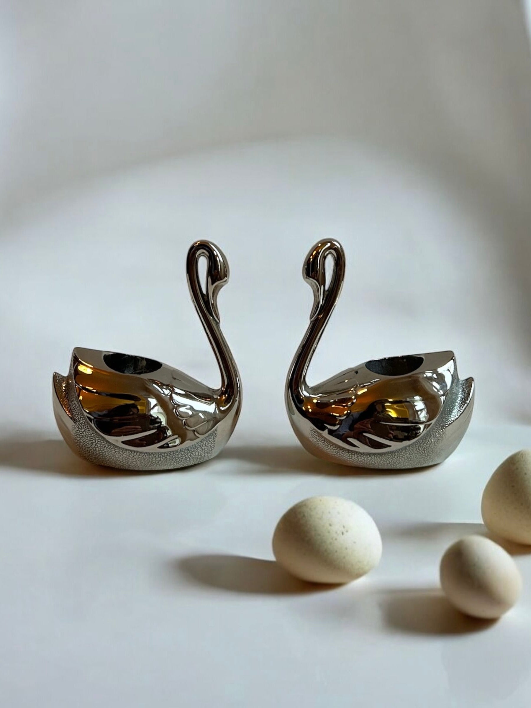 The Twin Swan Candle Holders by Flock Studio stand elegantly side by side on a smooth surface, with three small speckled egg-like stones placed in front, creating a sophisticated setting that is enhanced by the soft and blurred background.