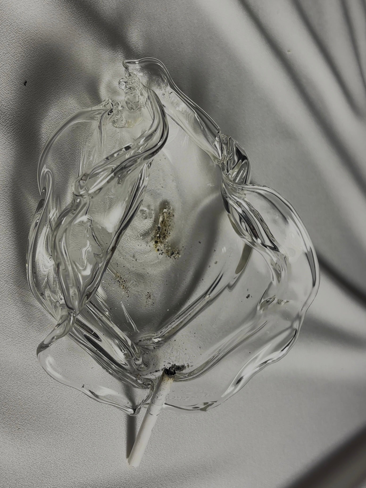 A top-down view of the Transparent Melted Ashtray by Agustina Ros, made of clear borosilicate glass, holding ashes and a partially burned cigarette on a light textured fabric surface with soft shadows.