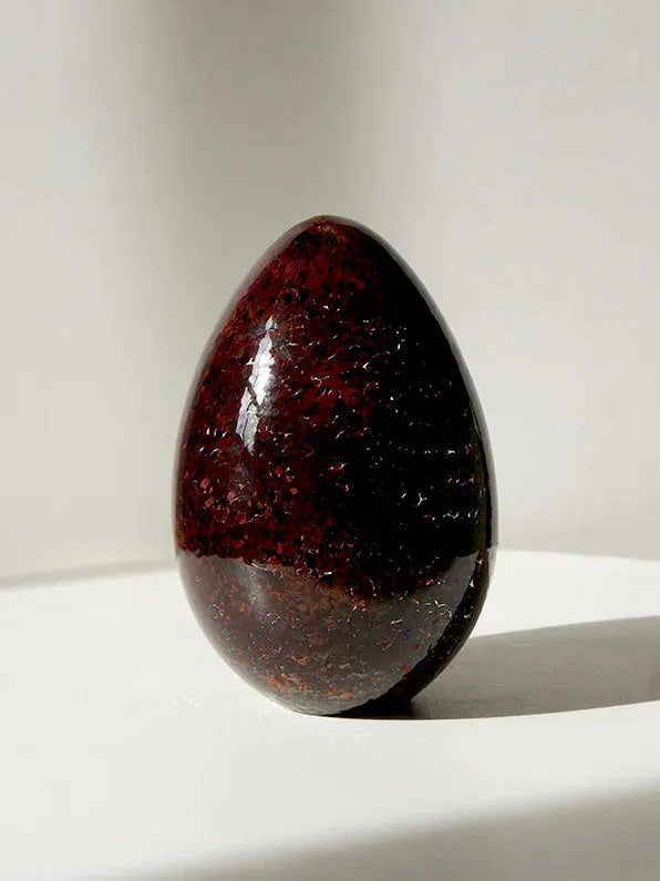 Close-up of the polished, dark reddish-brown Egg Paperweight by Vanderohe Curio, featuring a textured surface that mimics handblown craftsmanship. The paperweight is positioned on a smooth, white surface and brightly side-lit, casting a soft shadow. The background remains a soft and neutral white.