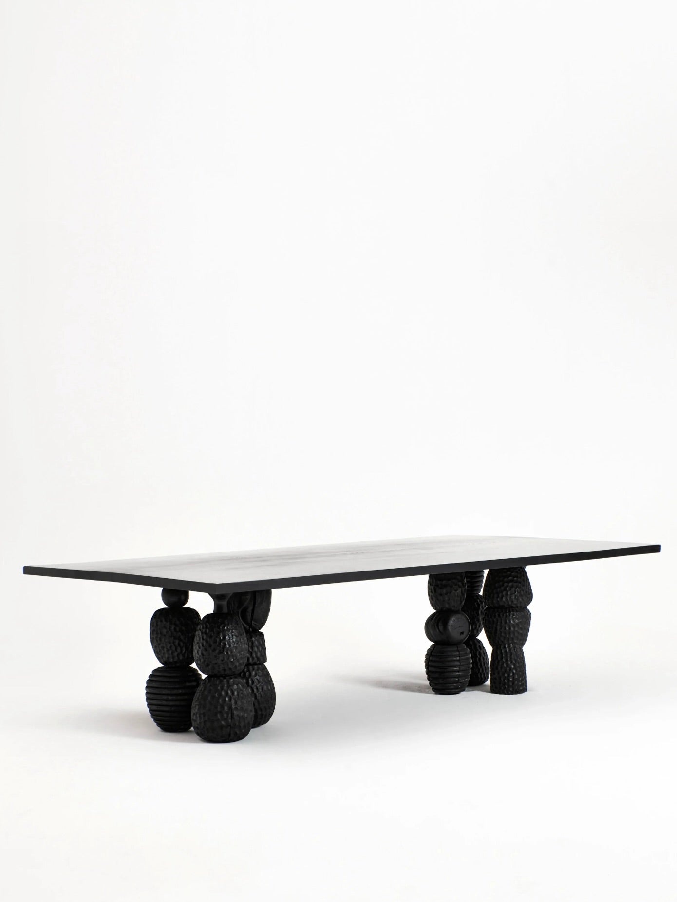 The Project 213A Menina Dining Table showcases a sleek black tabletop supported by four intricately sculpted legs. Handcrafted from chestnut wood, the legs feature an assortment of stacked black spheres and textured shapes, offering a striking modern aesthetic against a plain white backdrop.
