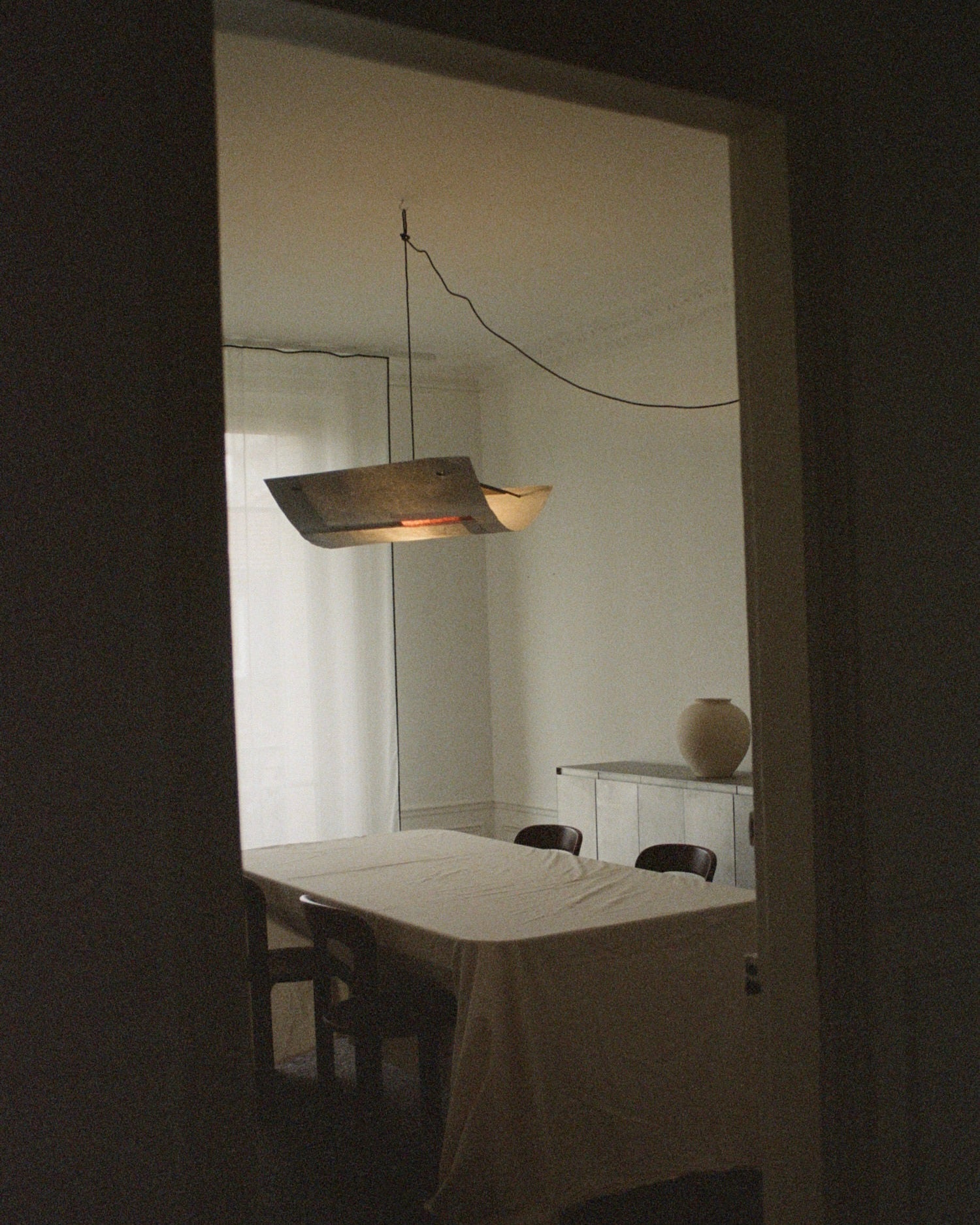 In the dimly lit dining room, a table is draped in cloth and surrounded by chairs. Overhead, a TEGET Sail Pendant Light – Lava gently illuminates the space. On the sideboard, a large vase sits against sheer curtains that softly filter light through.