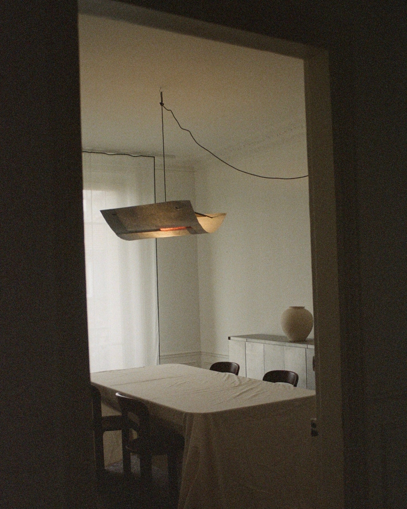 In the dimly lit dining room, a table is draped in cloth and surrounded by chairs. Overhead, a TEGET Sail Pendant Light – Lava gently illuminates the space. On the sideboard, a large vase sits against sheer curtains that softly filter light through.