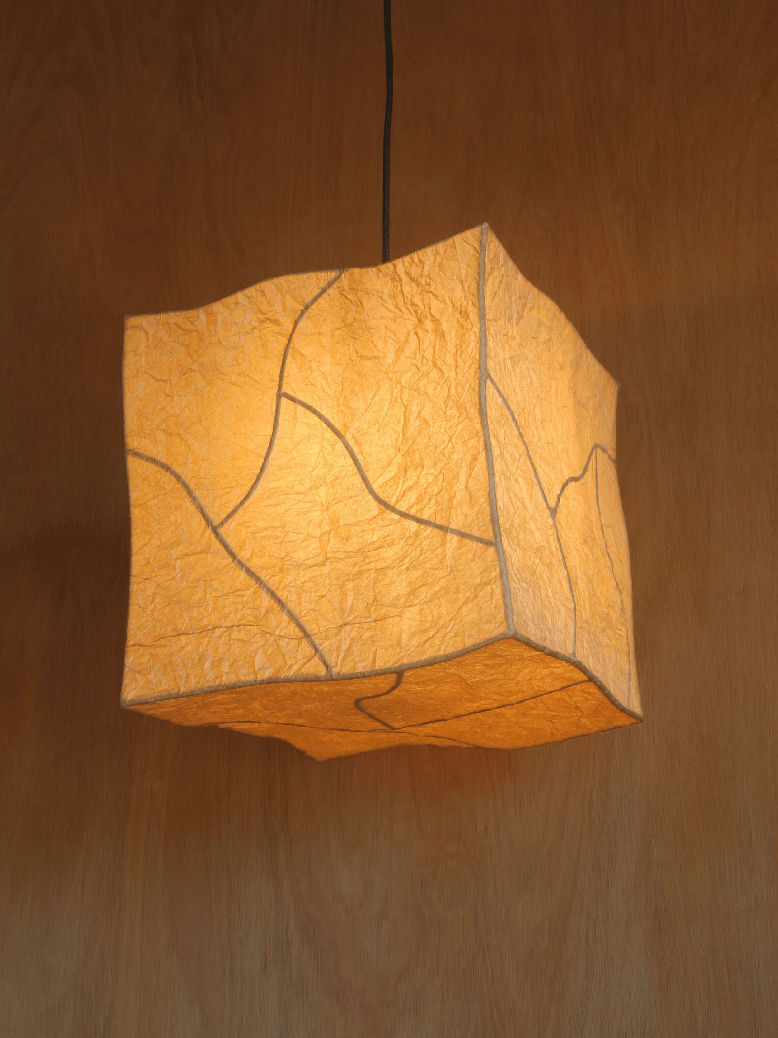 The Findere 'Hanging Dolmen' Pendant Light is a glowing, cube-shaped lamp made of textured crinkled paper with visible seams and natural oak bark dye, suspended from the ceiling against a wooden background.