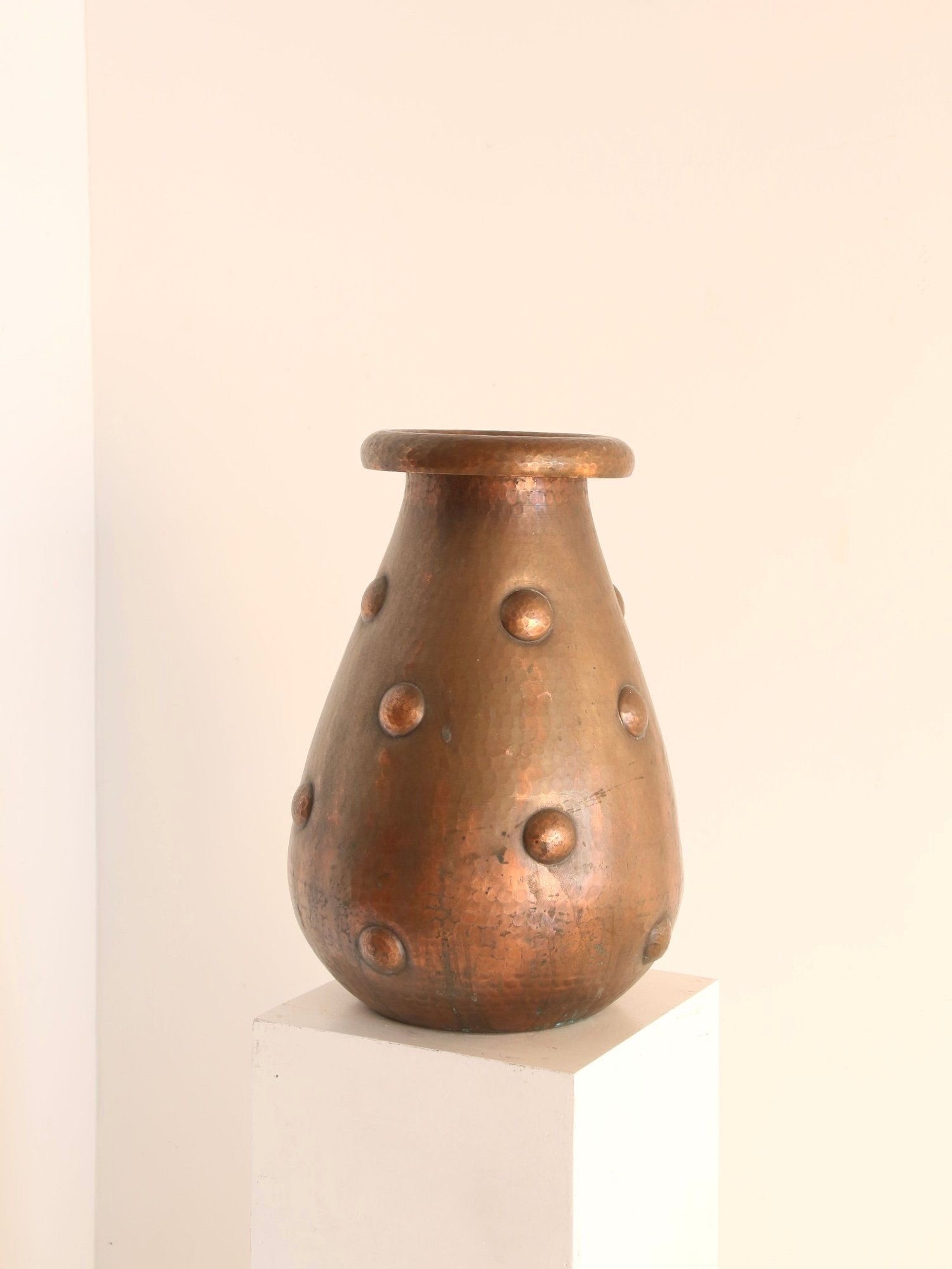 The Hand-Beaten Copper Vase by Ancien et Jolie, with its pear-shaped design and hammered texture showcasing raised circular motifs, embodies the Arts and Crafts spirit on a white pedestal against a plain beige background.