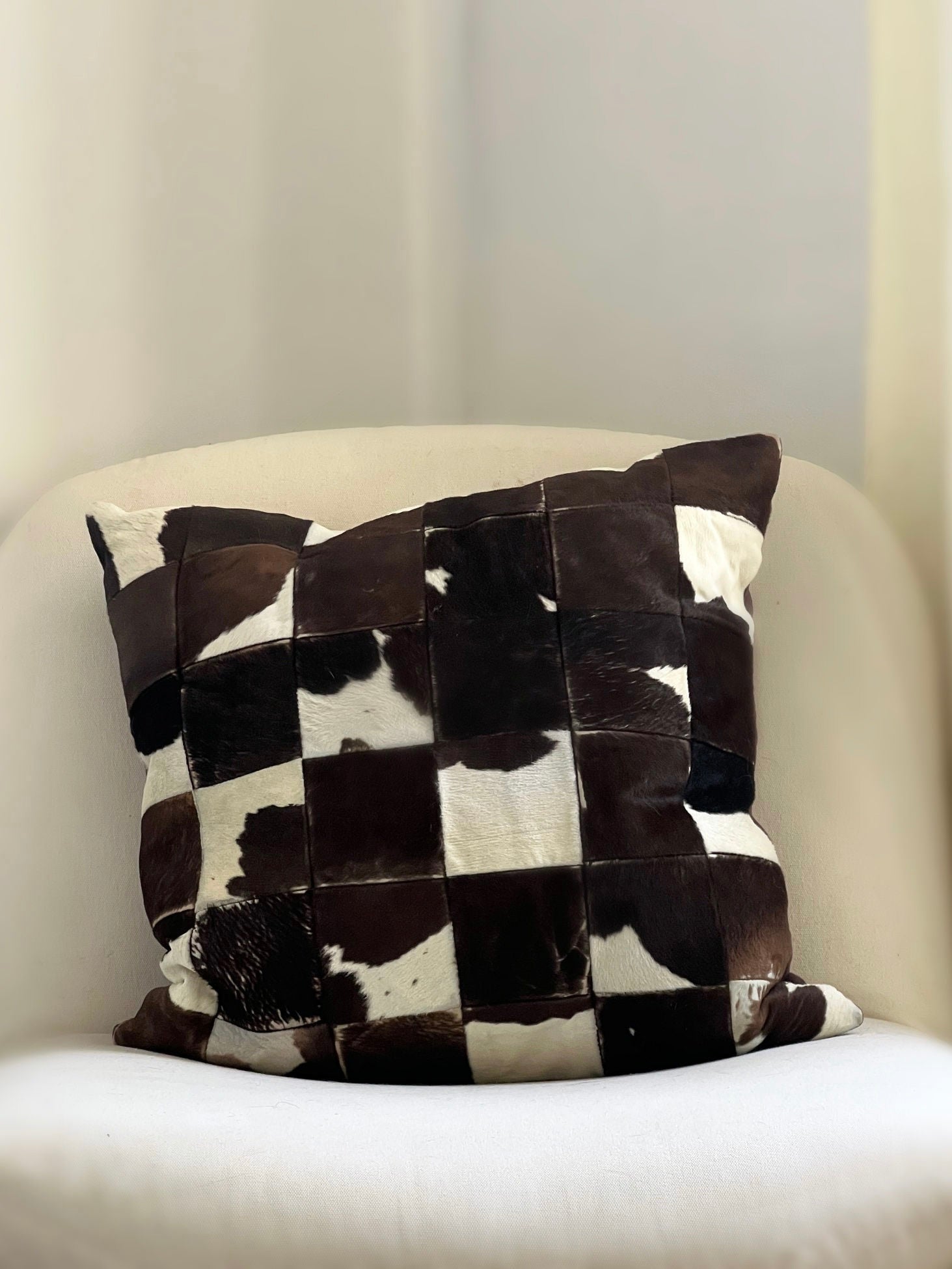The Porter Studio Cowhide Patchwork Cushion, featuring black, white, and brown cowhide designs and soft duck feather filling, sits on a cream armchair with light curtains as the backdrop.