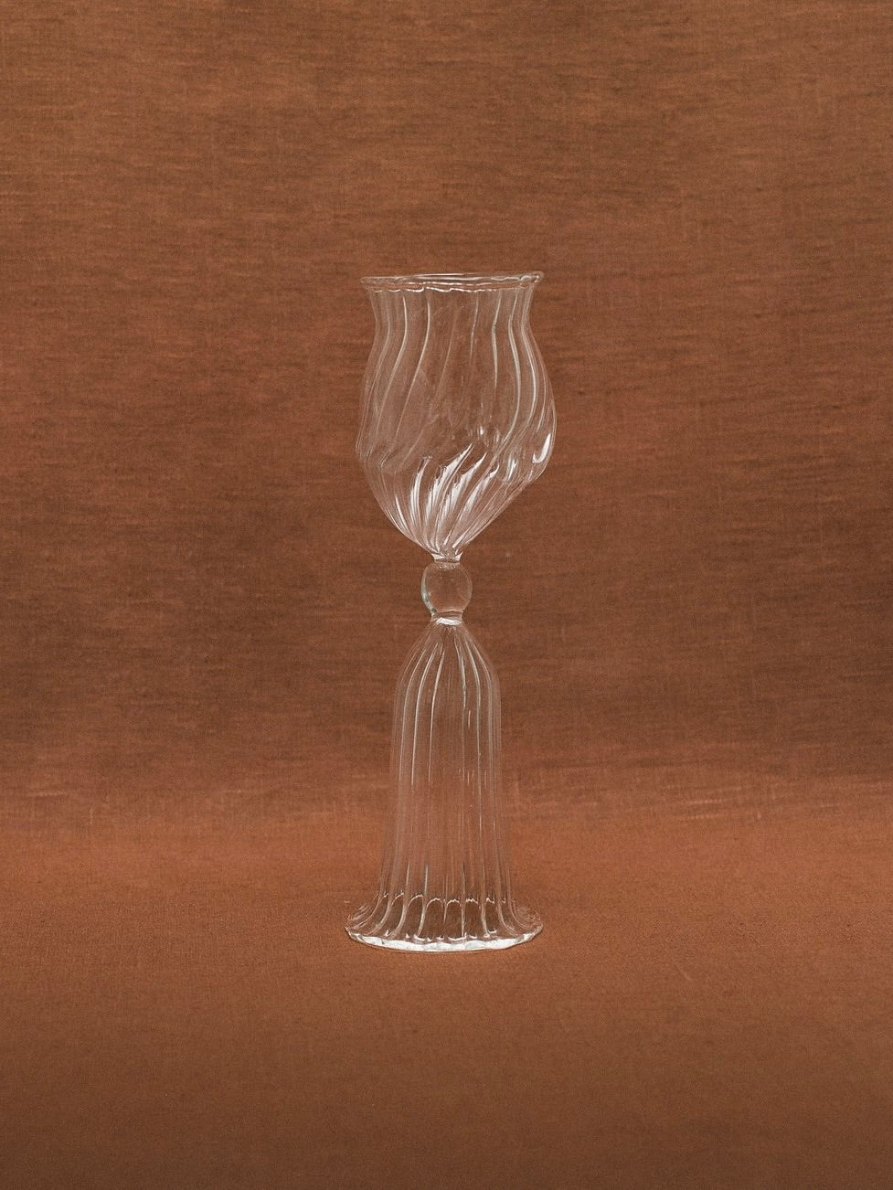 A mouth-blown glass goblet from the Cléodore Glass - Set Of Two by Justine Menard, with a transparent, swirled design and an ornate stem, is centered against a brown, textured background. The glass features intricate ridges and a distinctive twist in its design, creating an elegant and artistic appearance perfect for classic home decor.