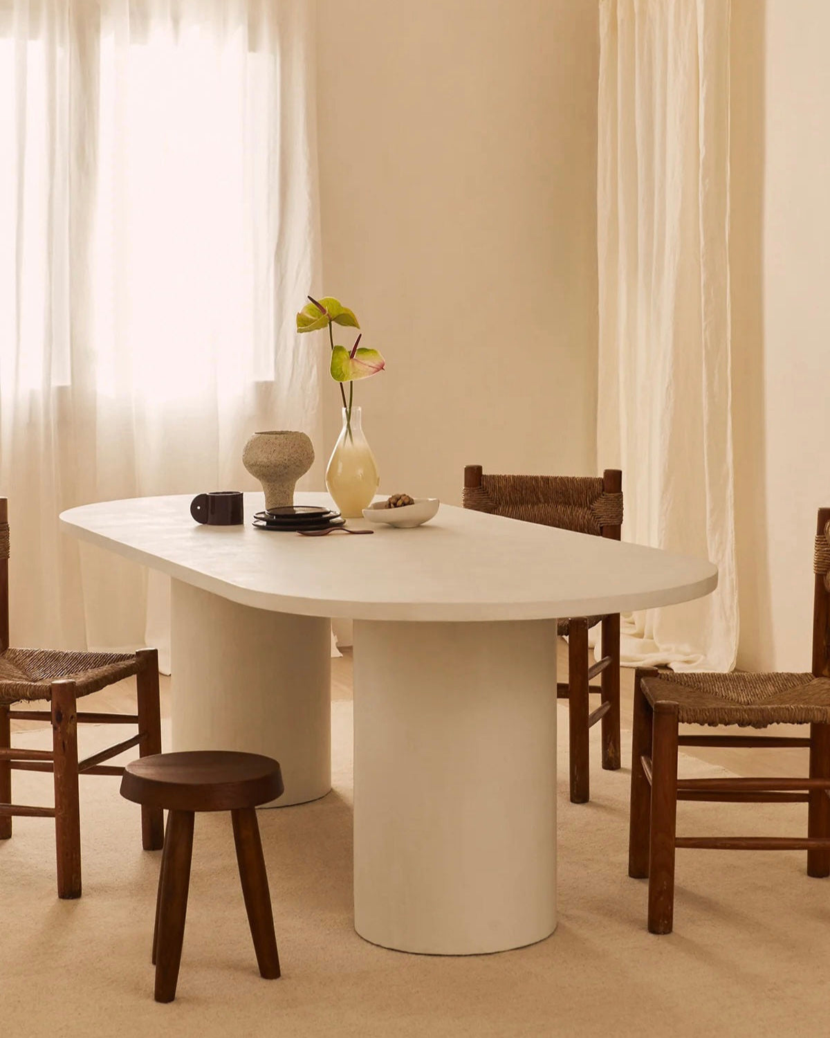 A minimalist dining room highlights the MarlotBaus Reye Dining Table, accompanied by wooden chairs and a stool. The table is adorned with a vase of flowers, a bowl, a mug, and decor. Soft natural light softly diffuses through sheer curtains.