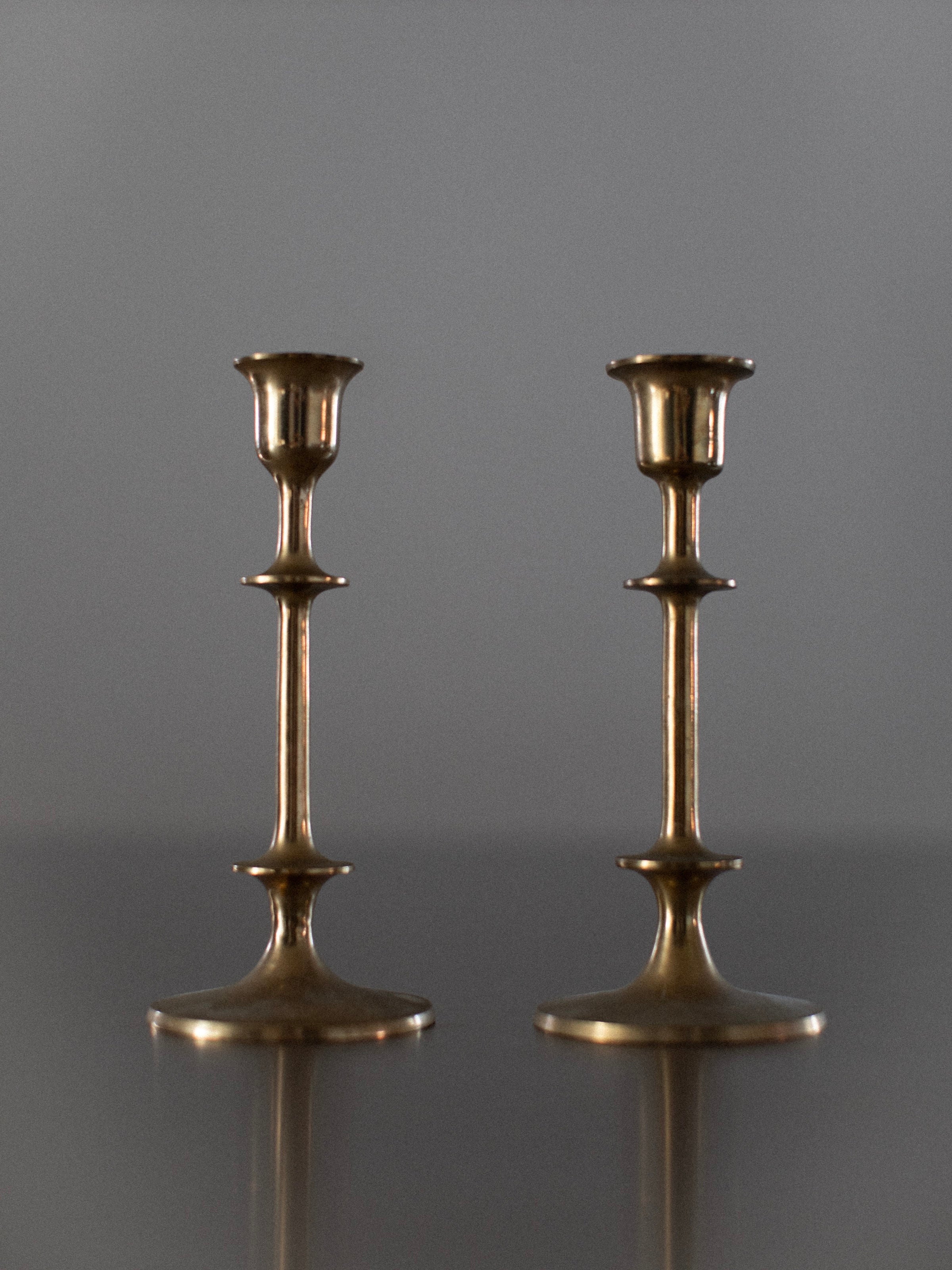 Two Out For Lunch Brass Candleholders stand upright on a flat surface against a plain, neutral background. Each candlestick boasts an elegant design with a simple, slender form, featuring a round base, a central knob, and a holder for the candle at the top.