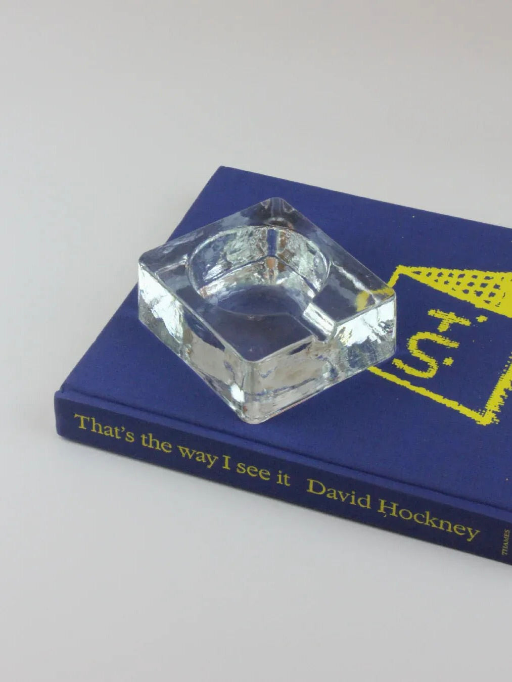 A Brutalist Ashtray from Boga Avante Shop, crafted in clear glass with a square shape, sits atop a blue book titled "That's the Way I See It" by David Hockney. The cover of the book also features a yellow abstract design, adding an artistic touch that complements the ashtray's style.
