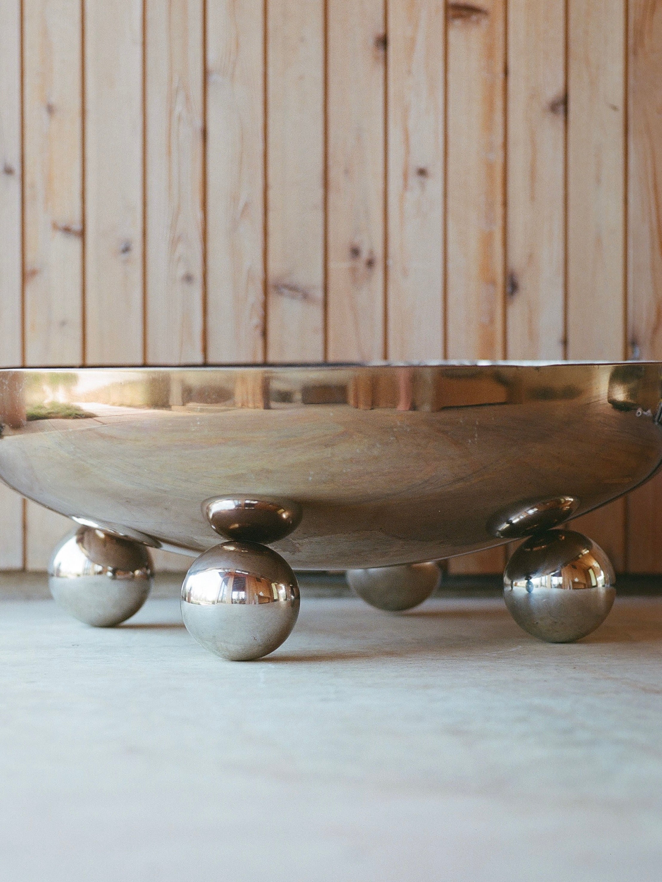 The MUHLY Flecto Fire Bowl, with its polished stainless steel surface, rests on a flat floor. This elegantly designed fire pit features six spherical legs for support and is positioned against a wooden-paneled wall backdrop. The reflective surface of the Flecto Fire Bowl reveals distorted reflections of the surroundings.