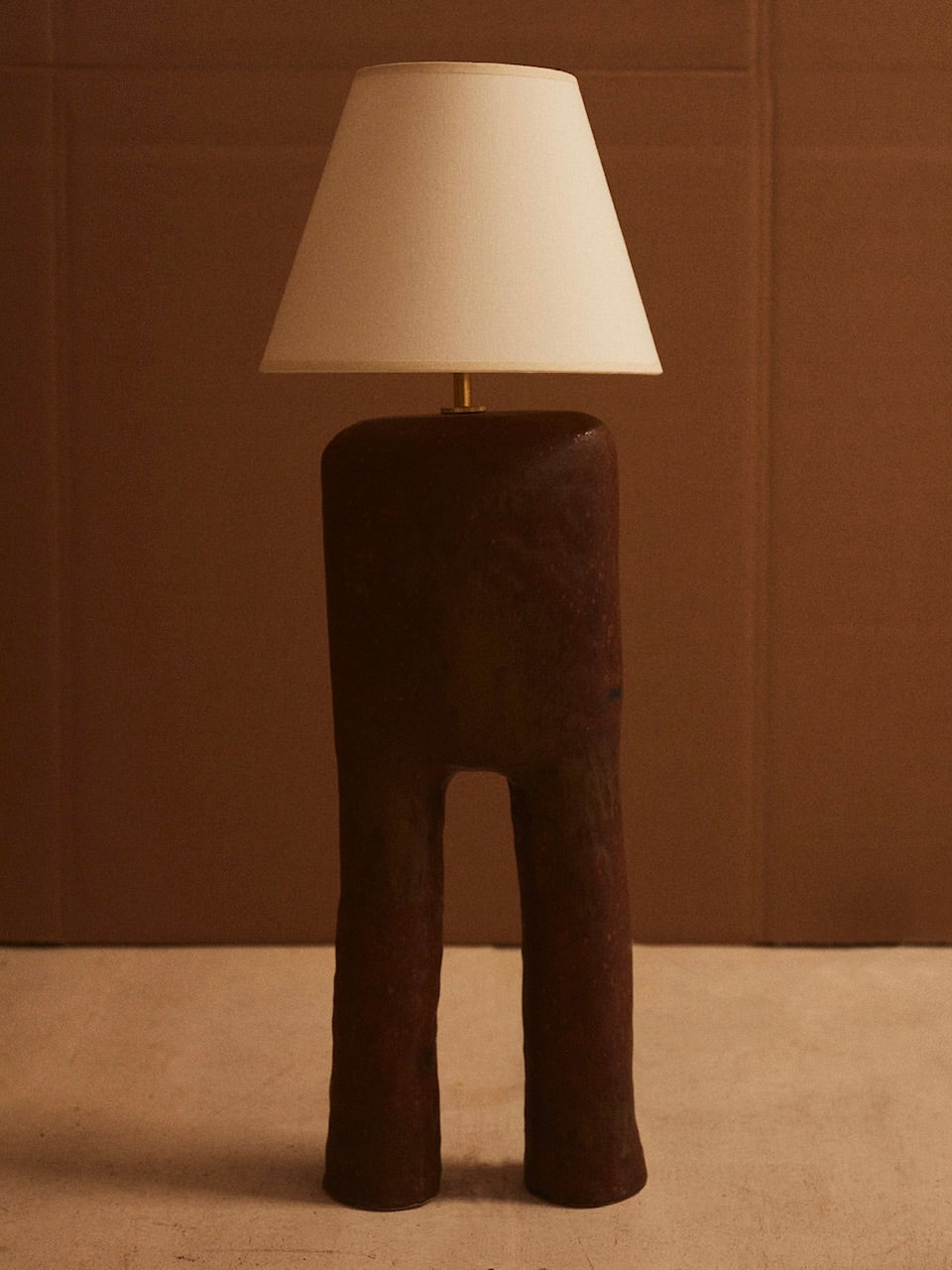 The LIV Table Lamp by marta bonilla features a unique base resembling abstract legs with a matte brown glaze, paired with a classic white lampshade. This design perfectly complements its neutral brown background, enhancing its rustic appearance.