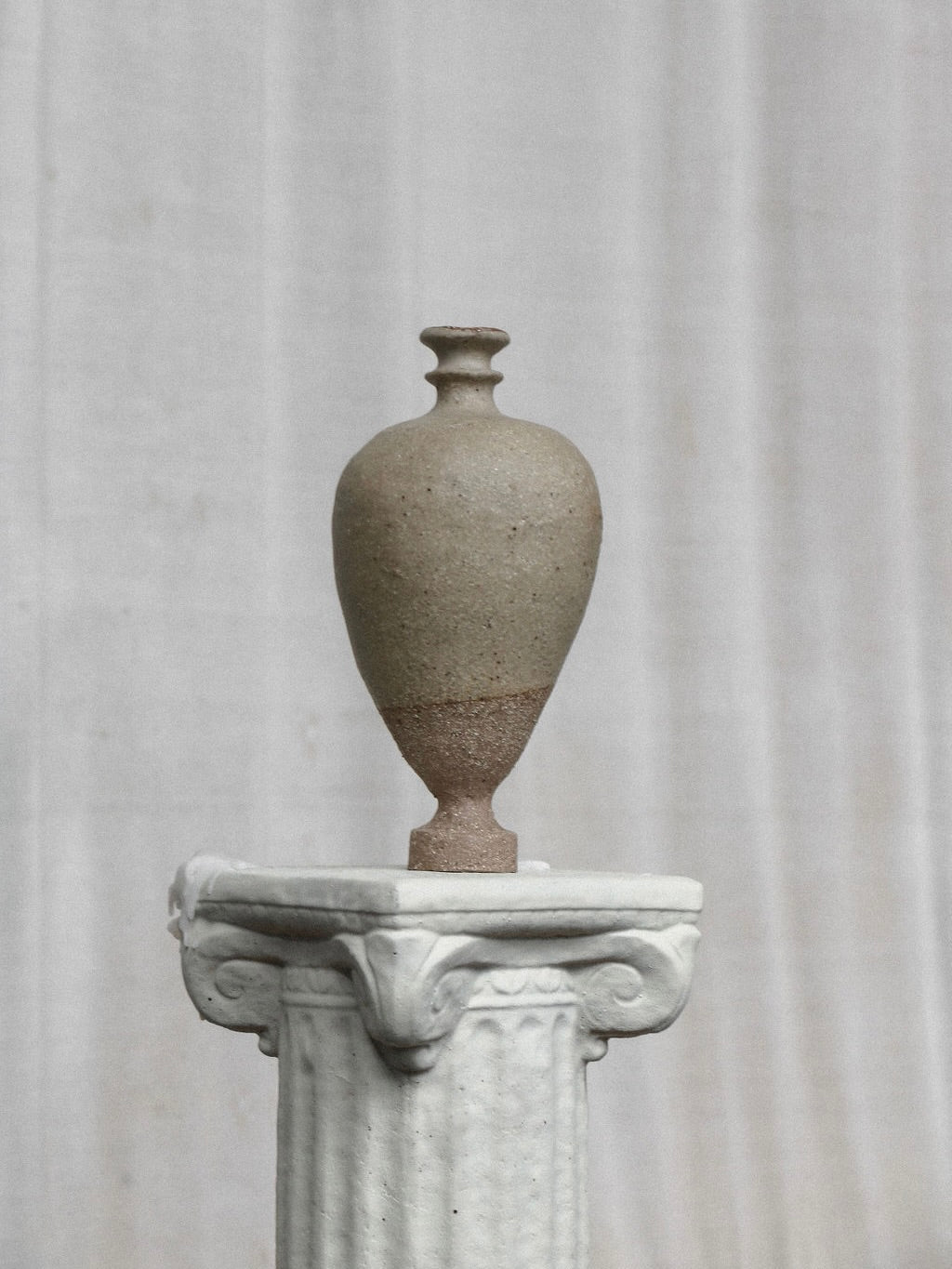 The Single Stem Vessel from Emma Lindegaard Studio, a tall, earthy-toned ceramic vase crafted by coiling, stands on a detailed white stone pedestal against a light gray backdrop. This hand-built piece features subtle texture and a narrow neck, showcasing its unique glaze-firing process.