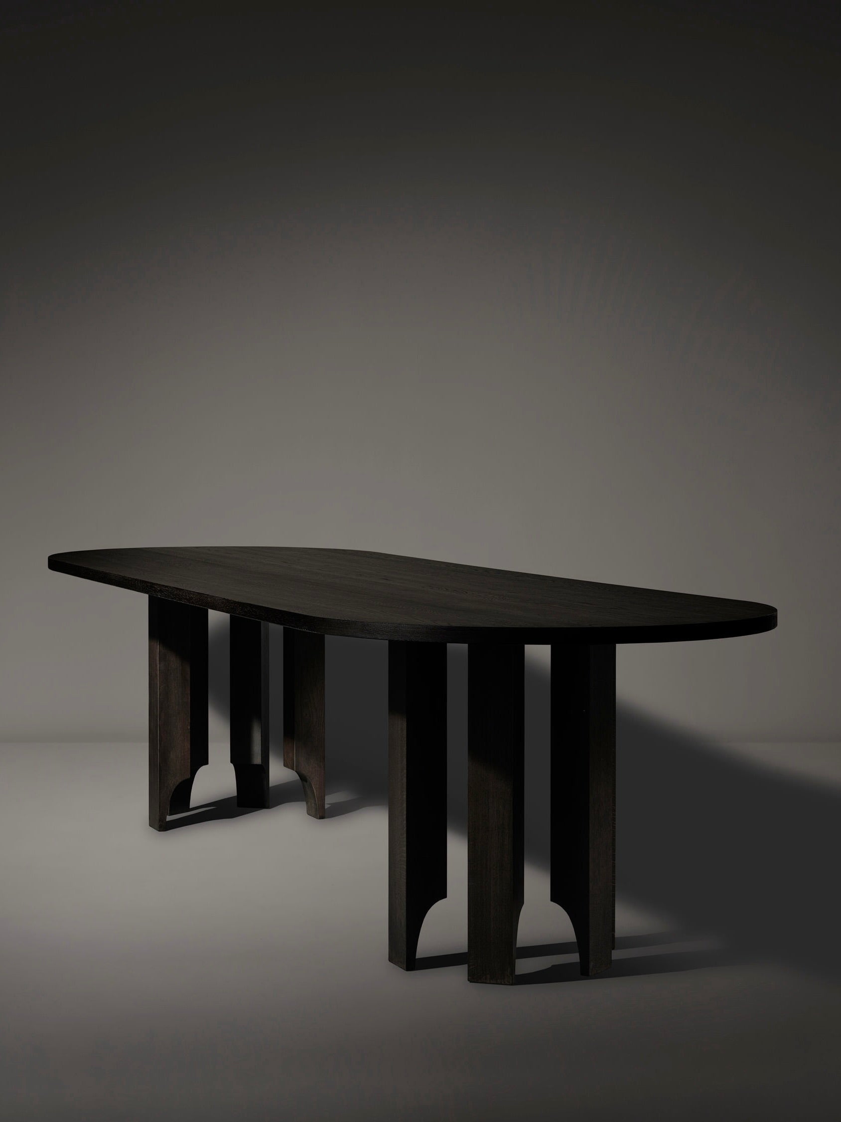 The Mokko Acer Capsule Table, crafted from solid European oak, features a smooth surface and unique arch-shaped legs. Set against a dark background, it showcases a minimalist modern design with subtle floor shadows.