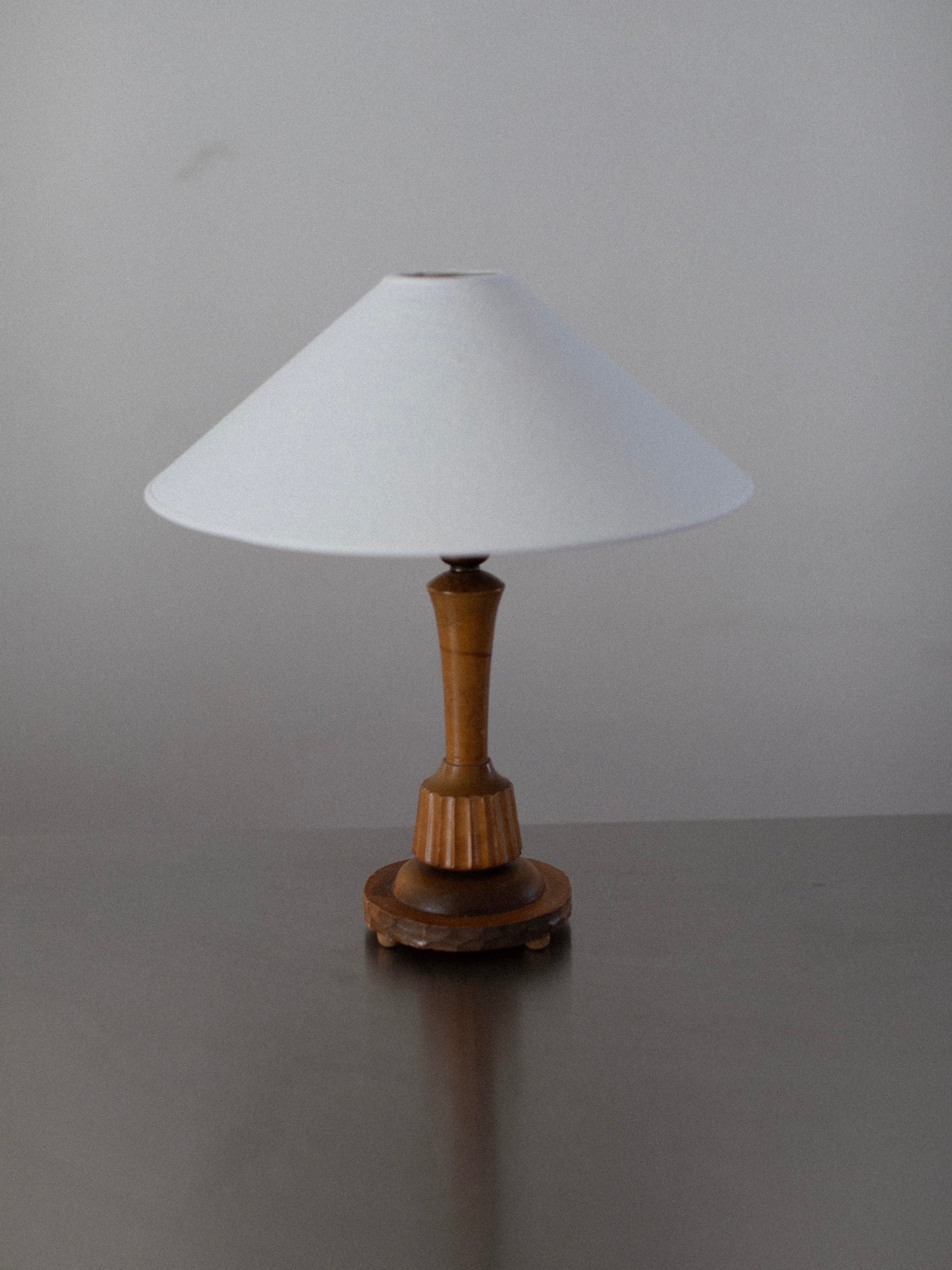 The Out For Lunch 1920's Table Lamp, featuring a vintage-inspired design with a white, cone-shaped lampshade and a hand-carved wooden base, is elegantly displayed on a gray surface against a plain gray background. This lamp beautifully captures the timeless charm of 1920's decor with its simple and classic design.