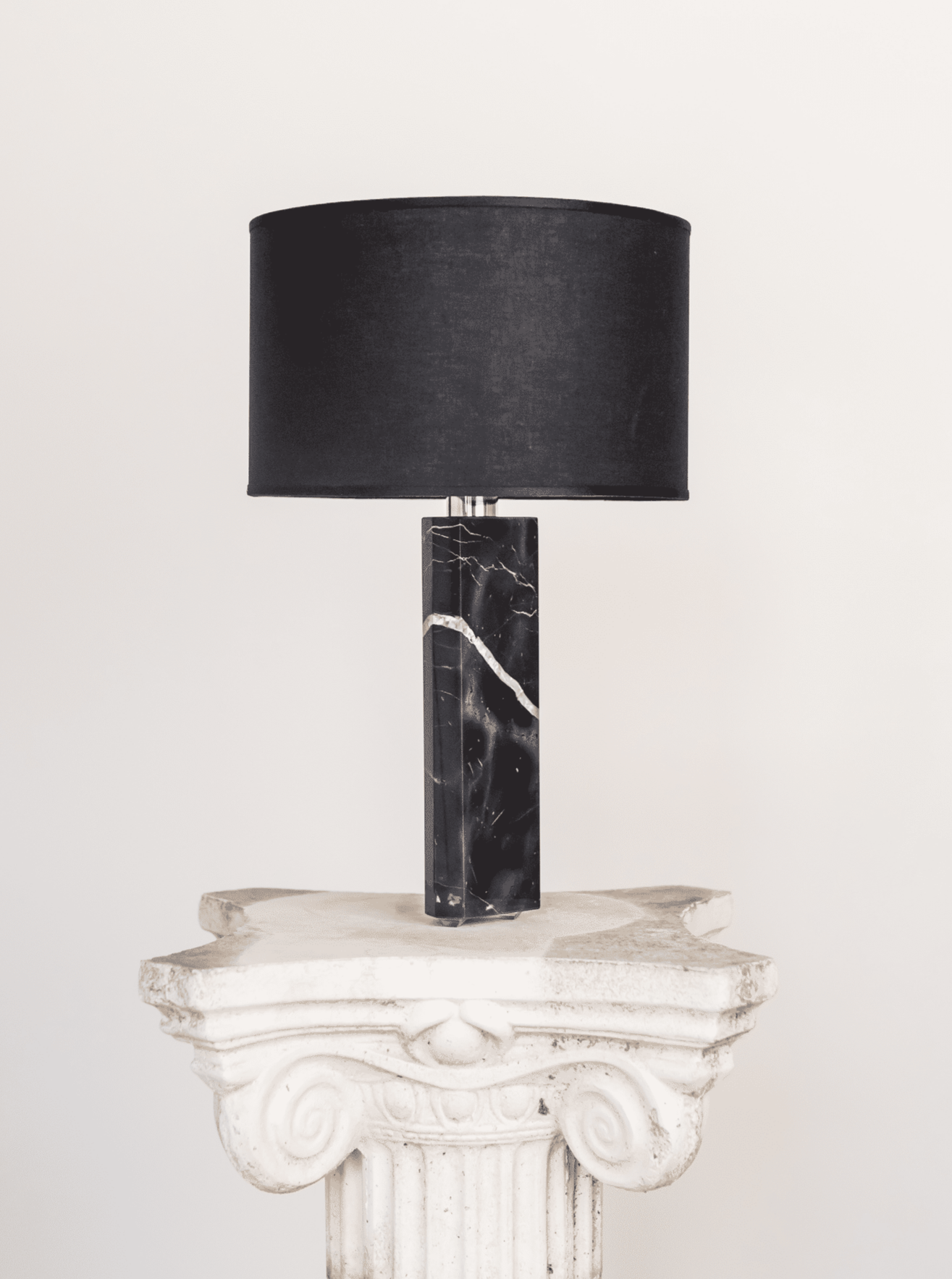 The Saint Laurent Table Lamp by Brajak Vitberg features a black cylindrical shade and a marble base with white veins. It sits on a decorative white pedestal with scrollwork, complementing luxury interiors against an off-white wall backdrop.
