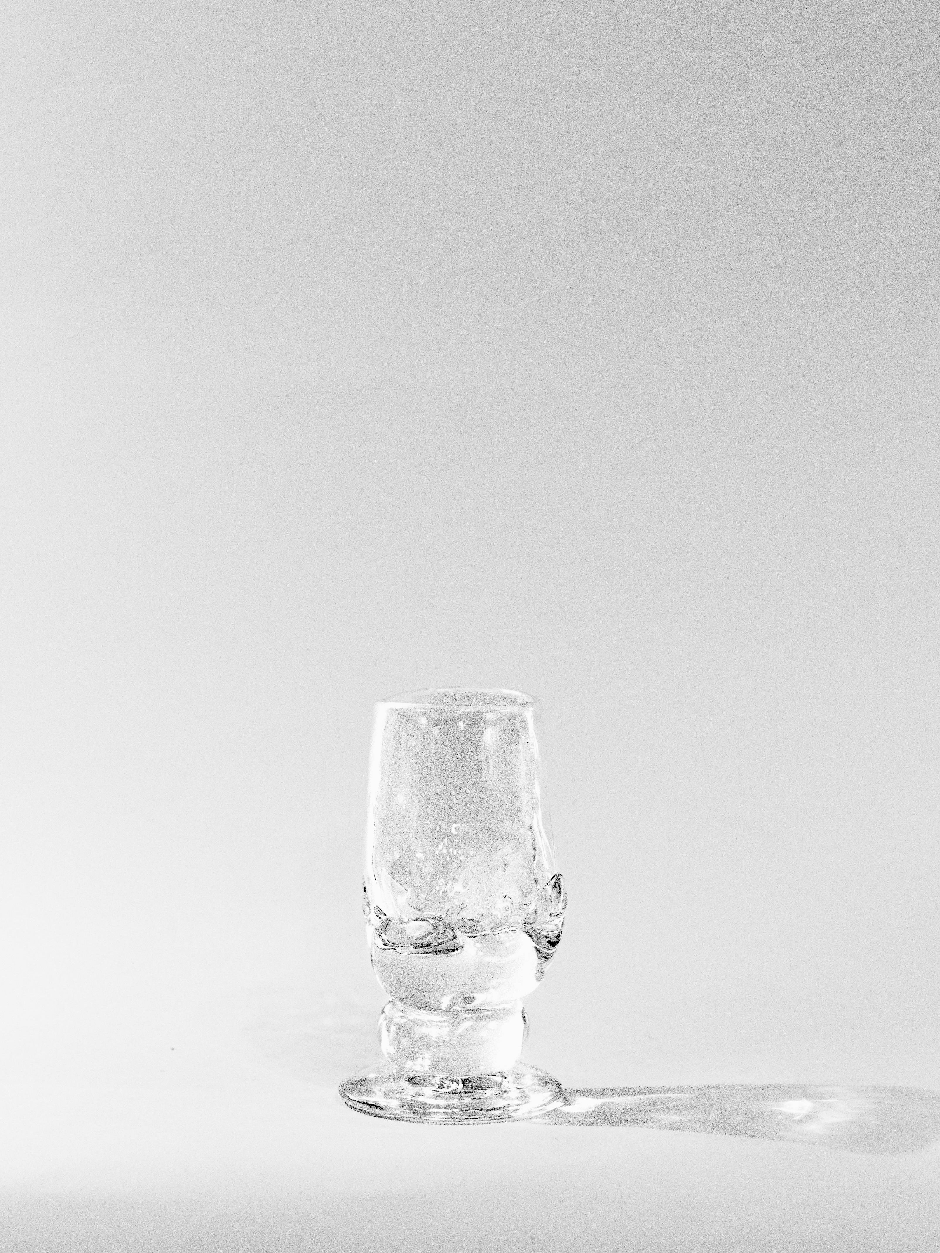 Against a plain white background, the Cocktail Goblet - Transparent from SzkĆo Studio, featuring a decorative base, stands centered. The lighting casts subtle shadows that highlight its freehand-blown contours and transparency.