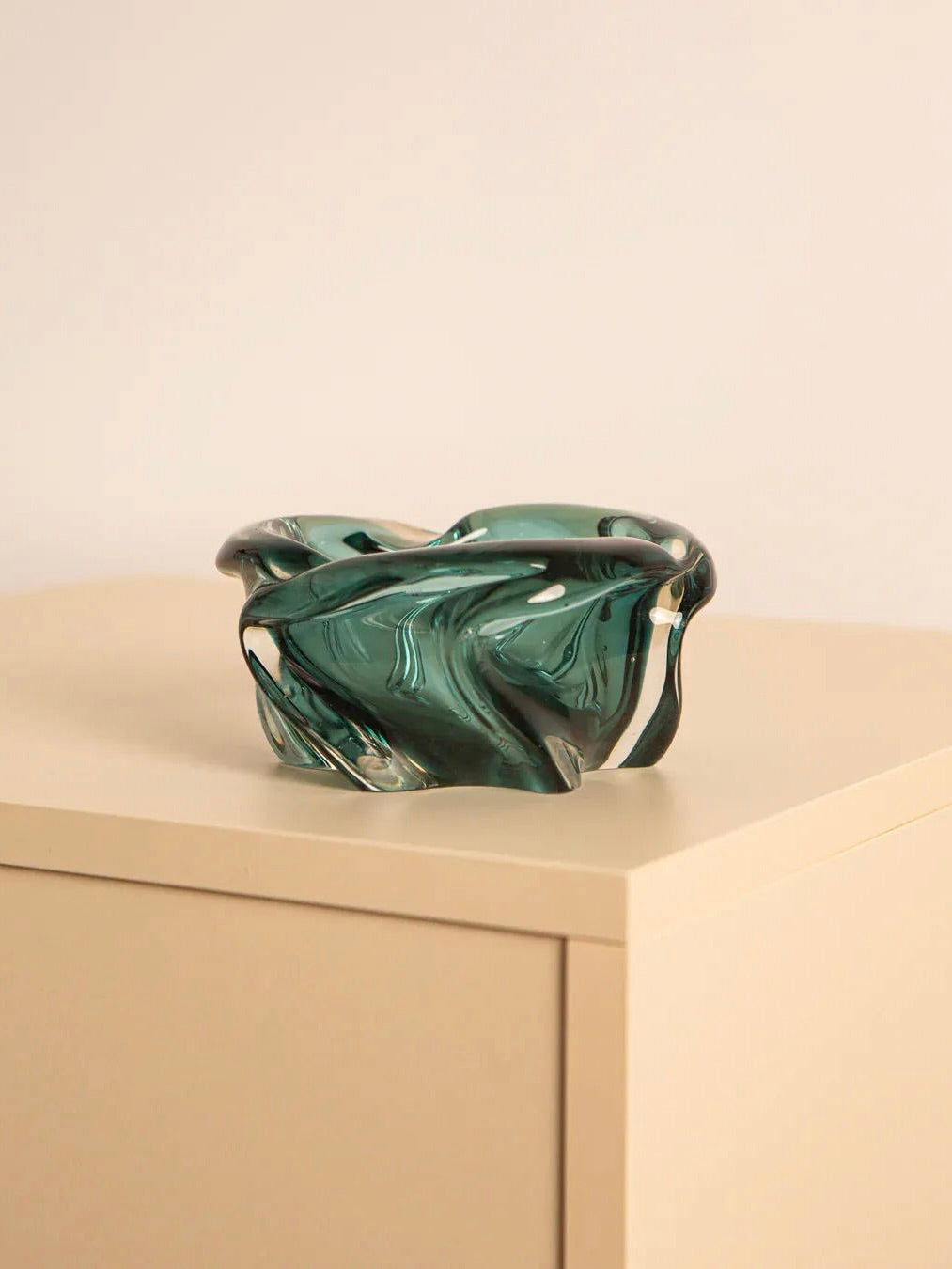 A Dark Turquoise Murano Glass Vide-poches by Treaptyque, featuring an abstract design, is placed on the edge of a beige square surface against a light gray backdrop.