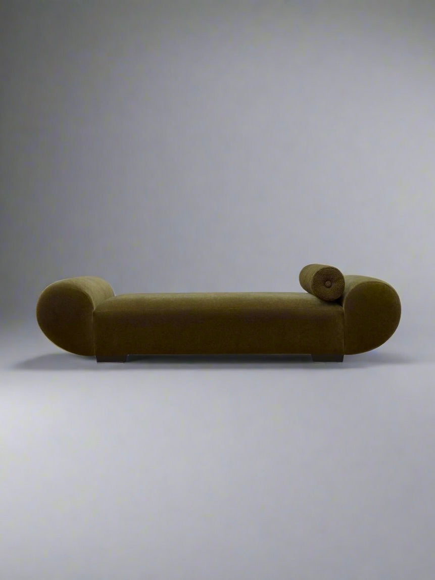 The "Sparta" Daybed by Ransom & Dunn, a modern olive-green chaise lounge featuring rounded armrests and a bolster cushion, is set against a plain light gray background. This minimalist piece, handmade in England, highlights clean lines and a streamlined silhouette.