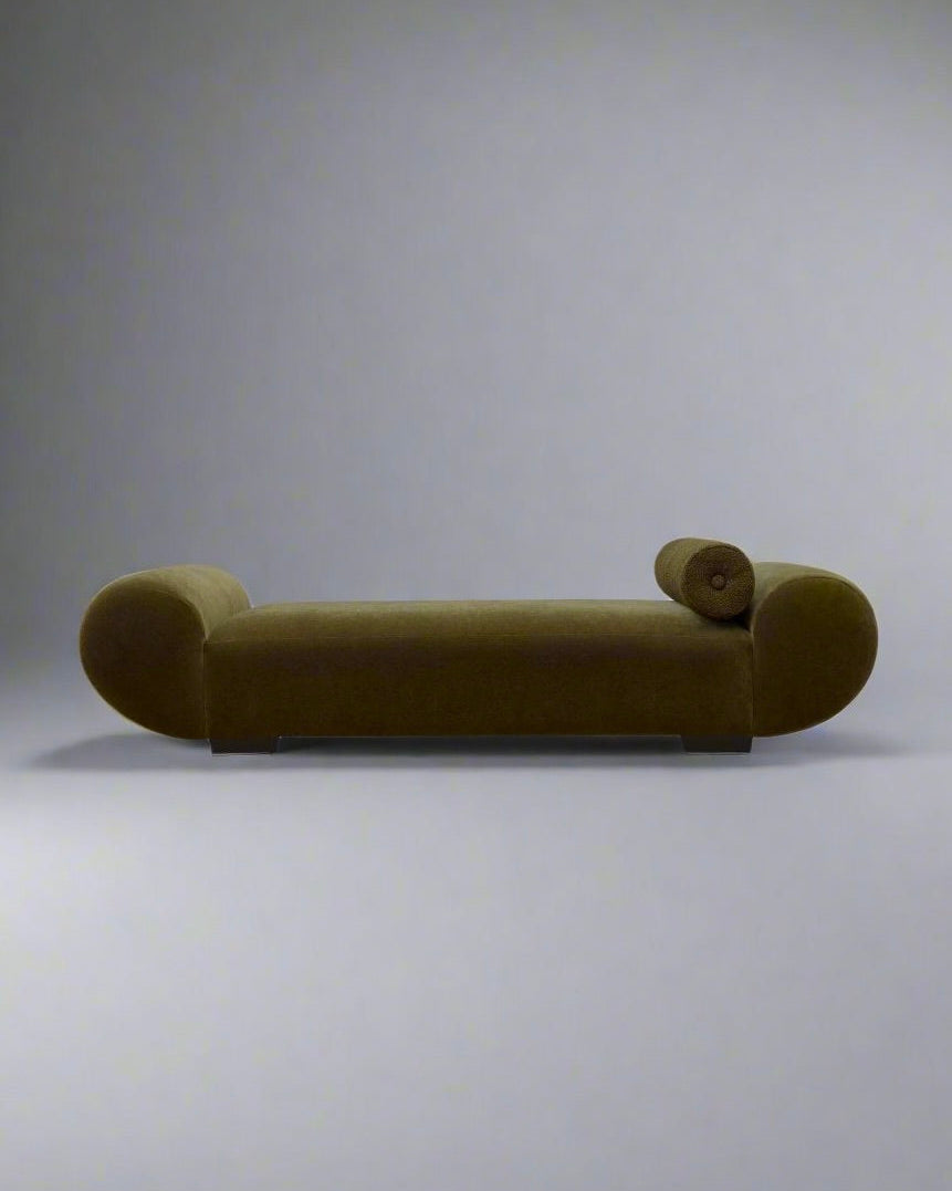 The "Sparta" Daybed by Ransom & Dunn, a modern olive-green chaise lounge featuring rounded armrests and a bolster cushion, is set against a plain light gray background. This minimalist piece, handmade in England, highlights clean lines and a streamlined silhouette.