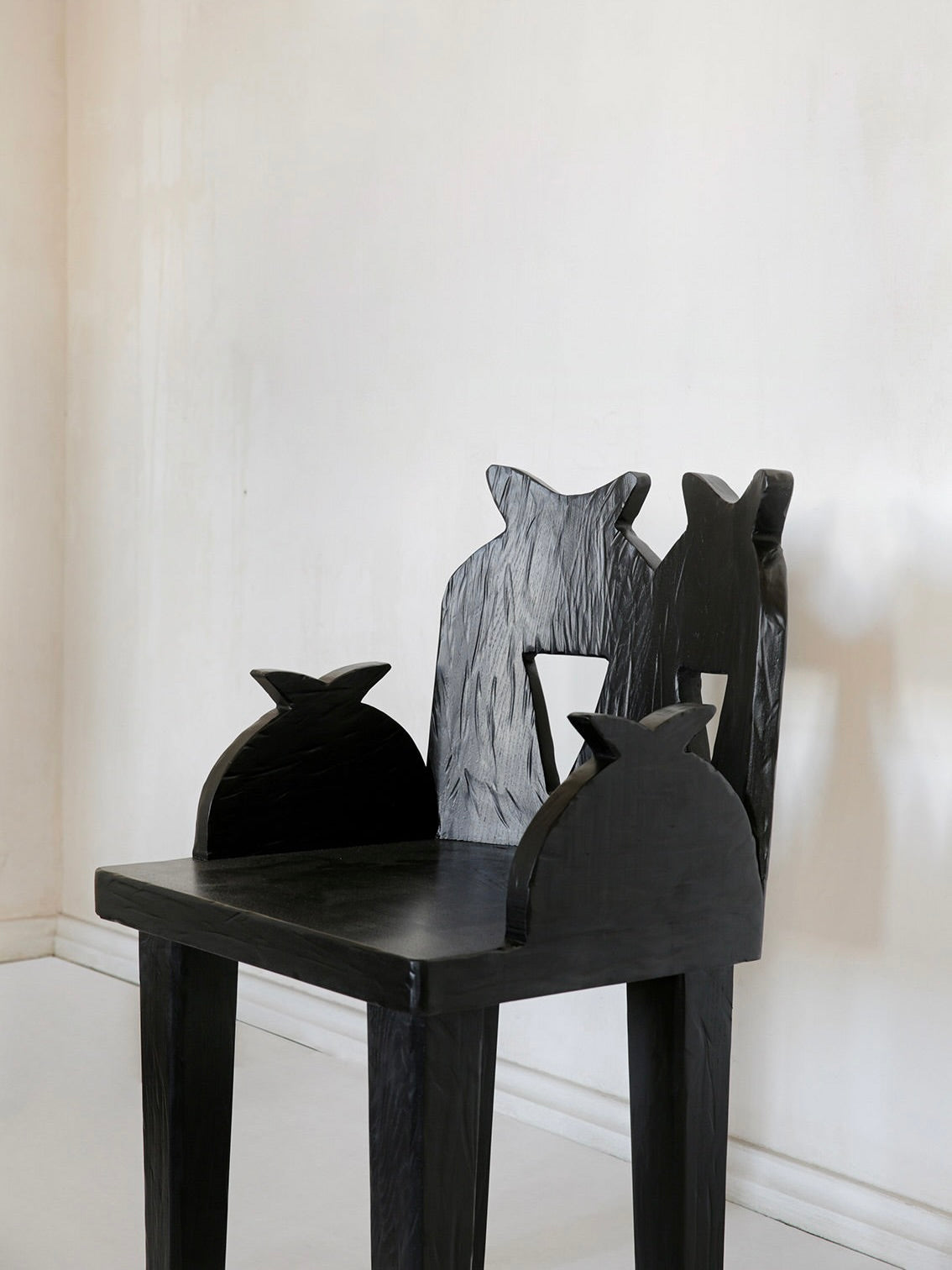 The Pomegranate Chair by Rooms Studio is a black reclaimed oak wooden chair with angular, jagged backrests and armrests resembling stylized animal silhouettes. It exudes contemporary flair and is set in a minimalistic, light-colored room.