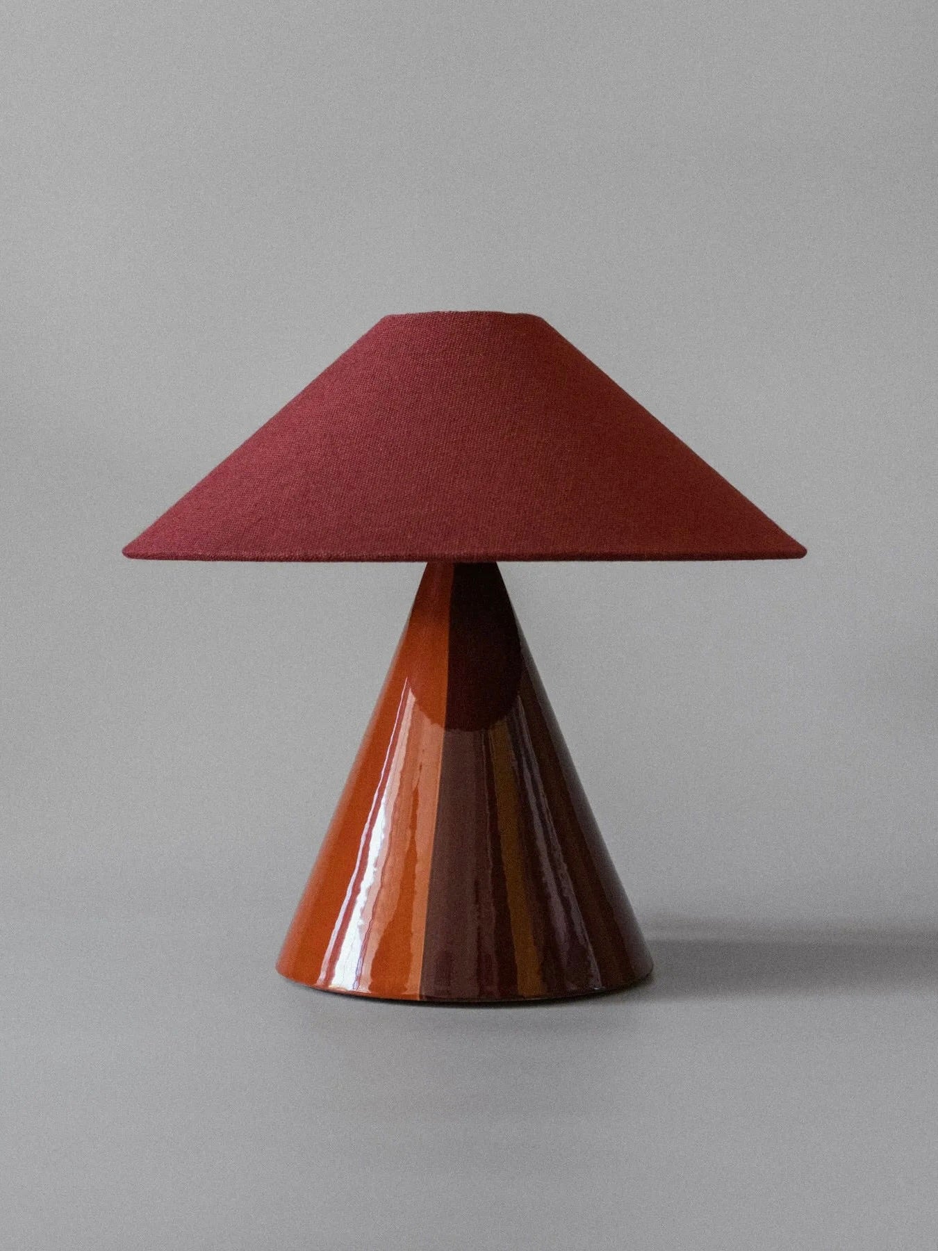The "Caterina" Table Lamp by mattina moderna boasts a conical, glossy brown earthenware base and a red, textured lampshade against a neutral gray background. This limited edition piece beautifully merges artistic design with contemporary charm.