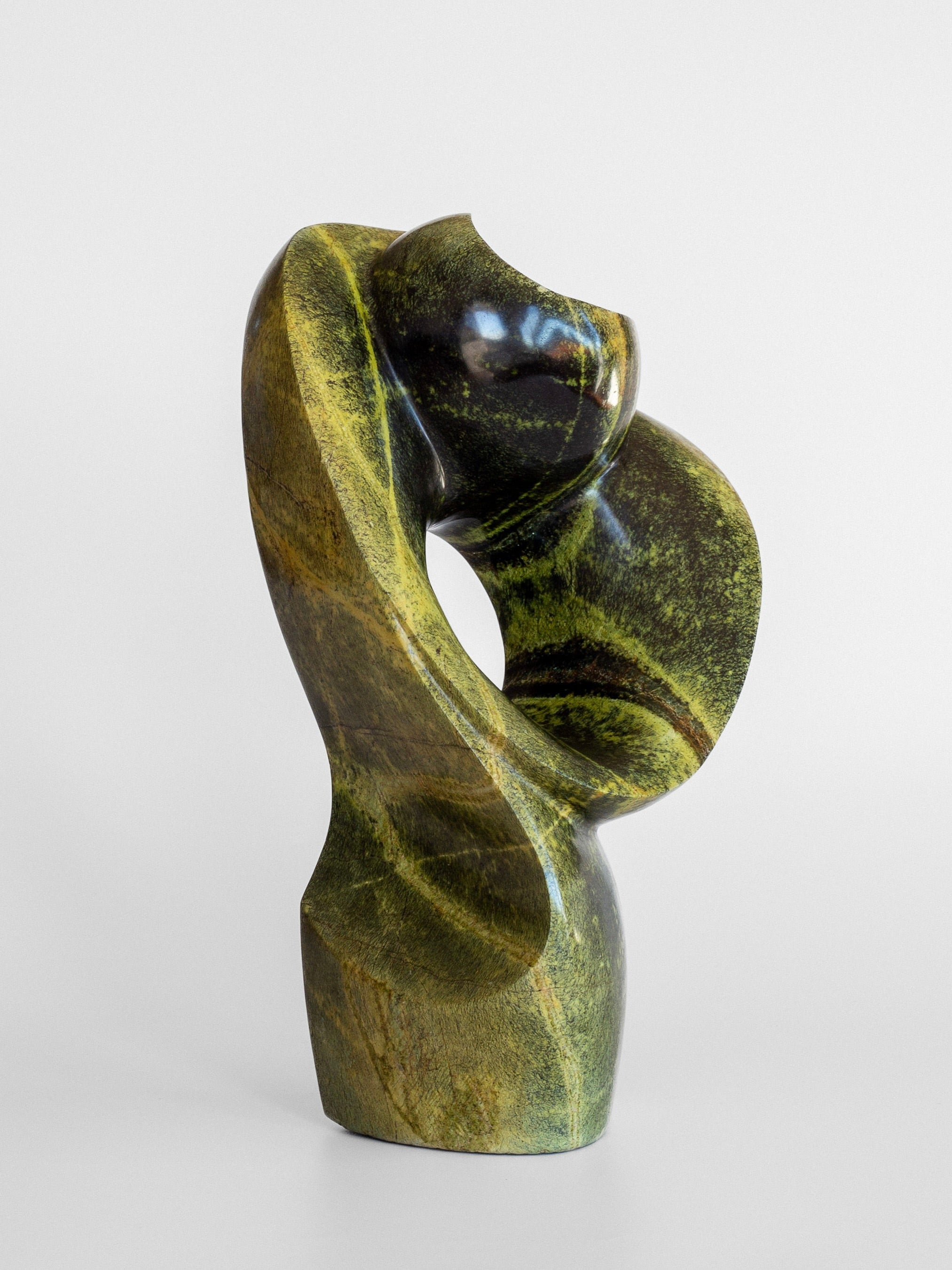 Presenting the "Abstract Stone Sculpture I" by Spigel, a stunning piece crafted by a skilled Zimbabwean artist from green and black leopard stone. This sophisticated sculpture features sleek curves and an elegant twist, creating a dynamic flow that conveys a sense of movement. Its polished surface subtly reflects light, enhancing its captivating aesthetic.