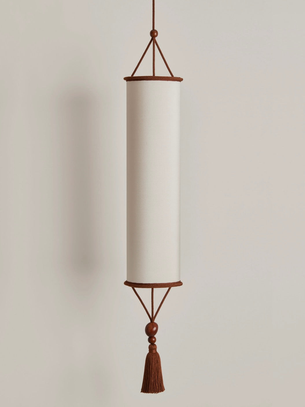 The Betty Pendant Lamp, designed by HAUVETTE & MADANI, is a minimalist cylindrical fixture with plant ivory brown trim and a tassel at the bottom, suspended by a matching cord against a light-colored wall.