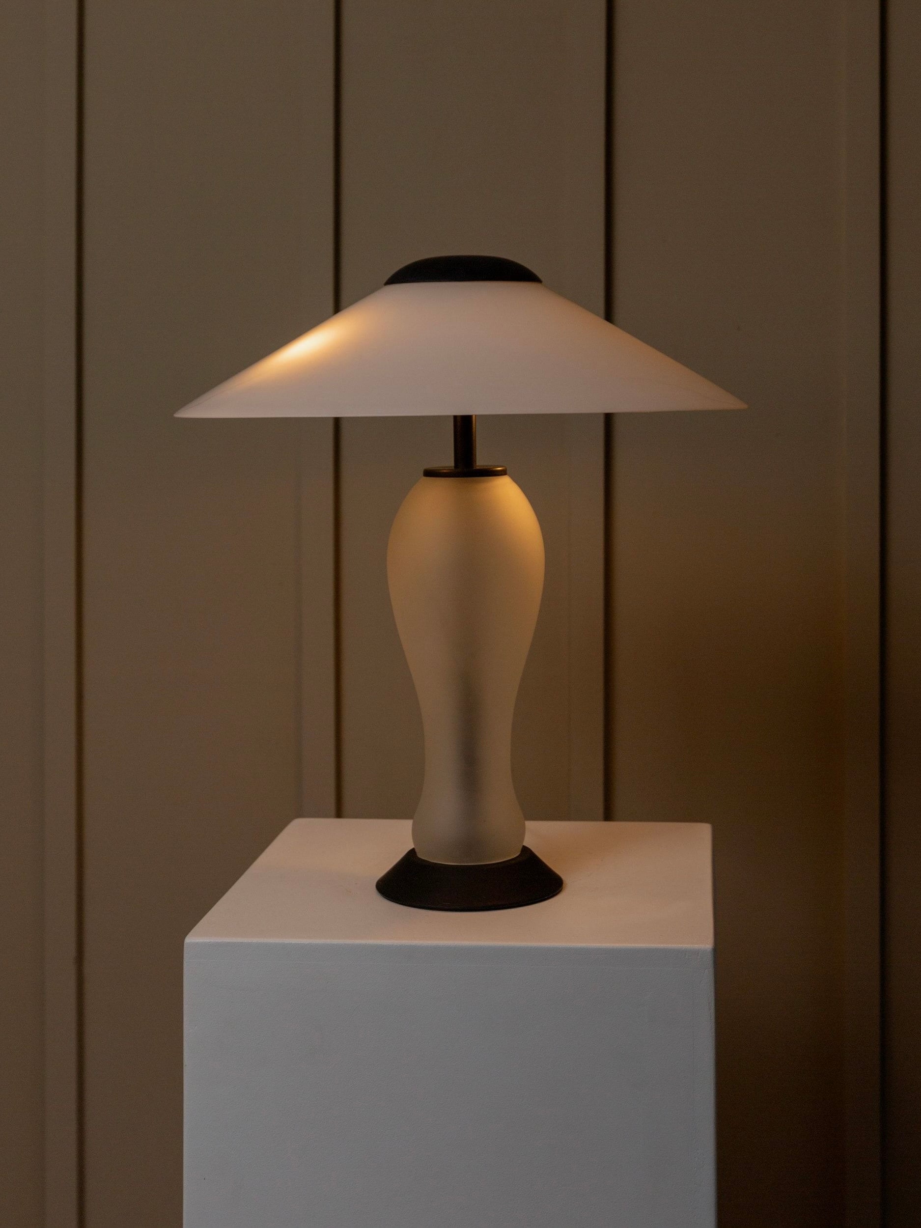The Vintage Murano Glass Lamp by Bottega Jacobs, in the classic 80s Memphis style, features a frosted glass base and a wide, conical lampshade that emits a soft, warm glow. Positioned on a rectangular white pedestal against a muted brown paneled wall, this elegant piece integrates effortlessly into any space.