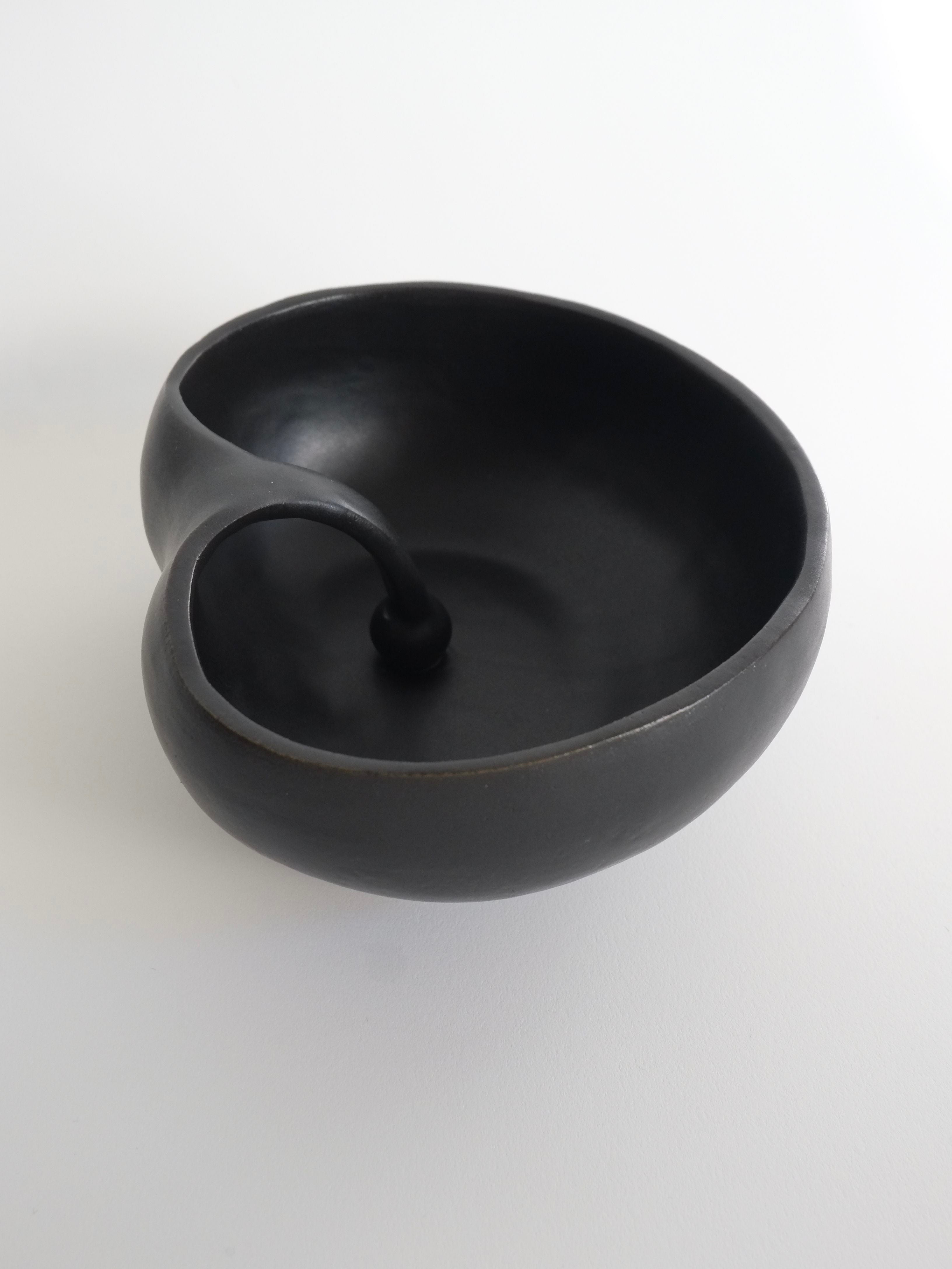 The Charlyn Reyes Seed Bowl, a handmade, uniquely shaped black ceramic piece with a looped handle, stands on a white background, showcasing its sculptural elegance.