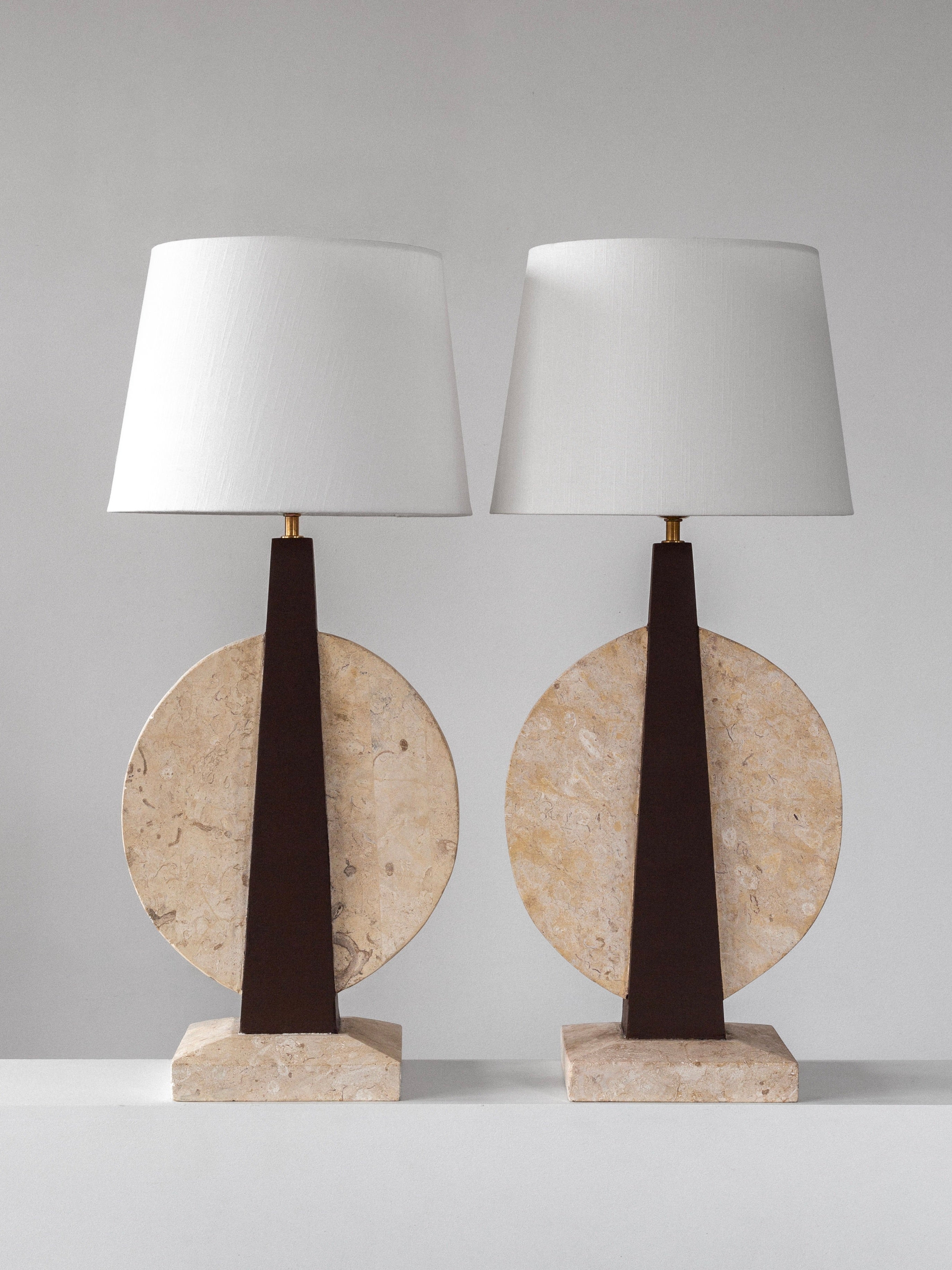 The Spigel Pair of Large Travertine Table Lamps from the 1970s features white cylindrical shades on triangular dark stands with circular stone bases atop rectangular platforms, symmetrically set against a plain backdrop to evoke vintage elegance.