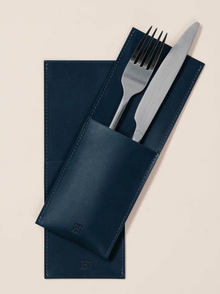 Paradise Row's Leather Cutlery Holder (set of 4) stylishly organizes cutlery including a knife and fork in a dark blue leather pouch on a light beige background, with the holder partially open to reveal utensil handles.