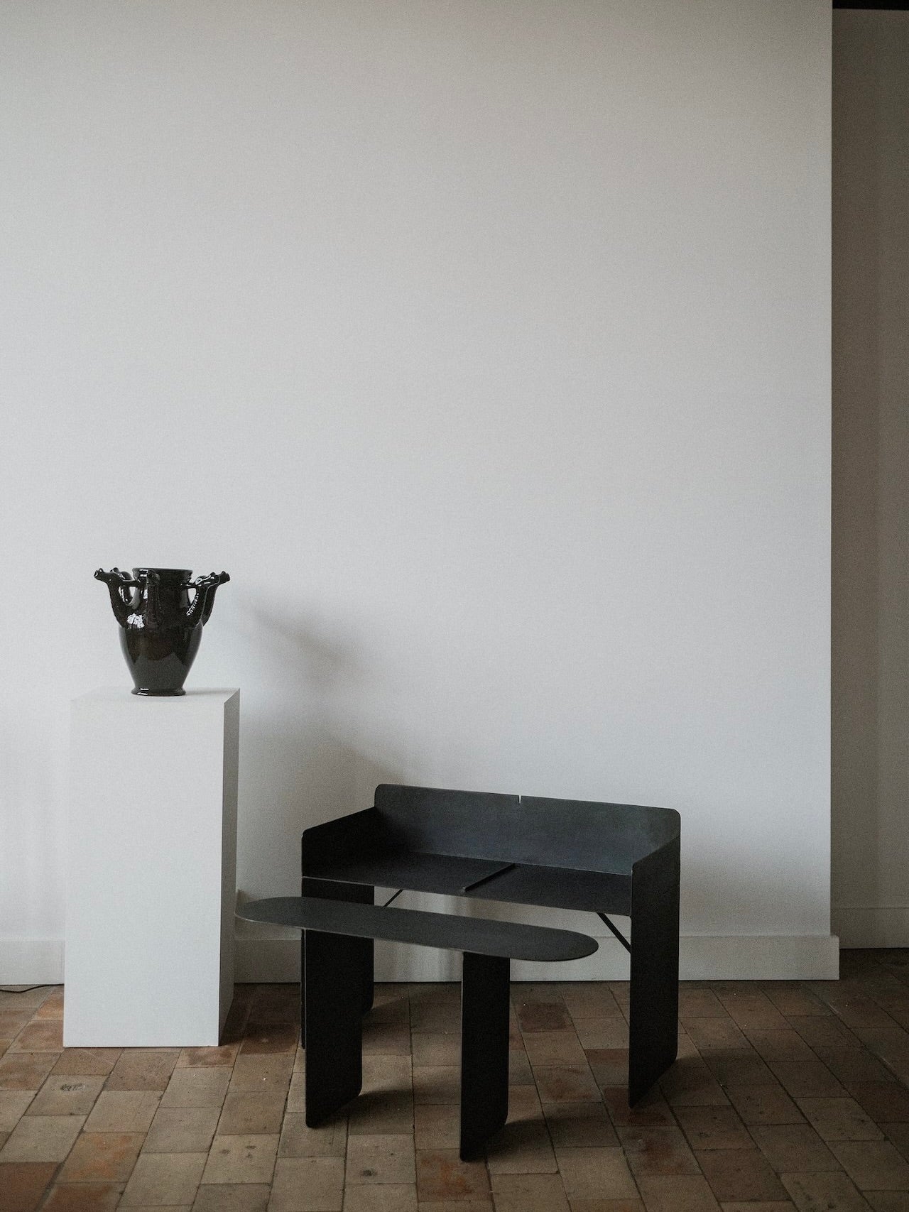 A minimalist room has a dark, modern chair on light wood flooring. To the left, a white pedestal displays a glossy black vase. A COSEINCORSO limited edition artisan handcrafted wooden bench complements the plain white wall in the background.