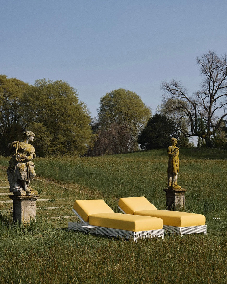 Two 'Conservatory' Sun Loungers by Lemon with yellow cushions sit on grass between two weathered stone statues, framed by large trees under a clear blue sky.