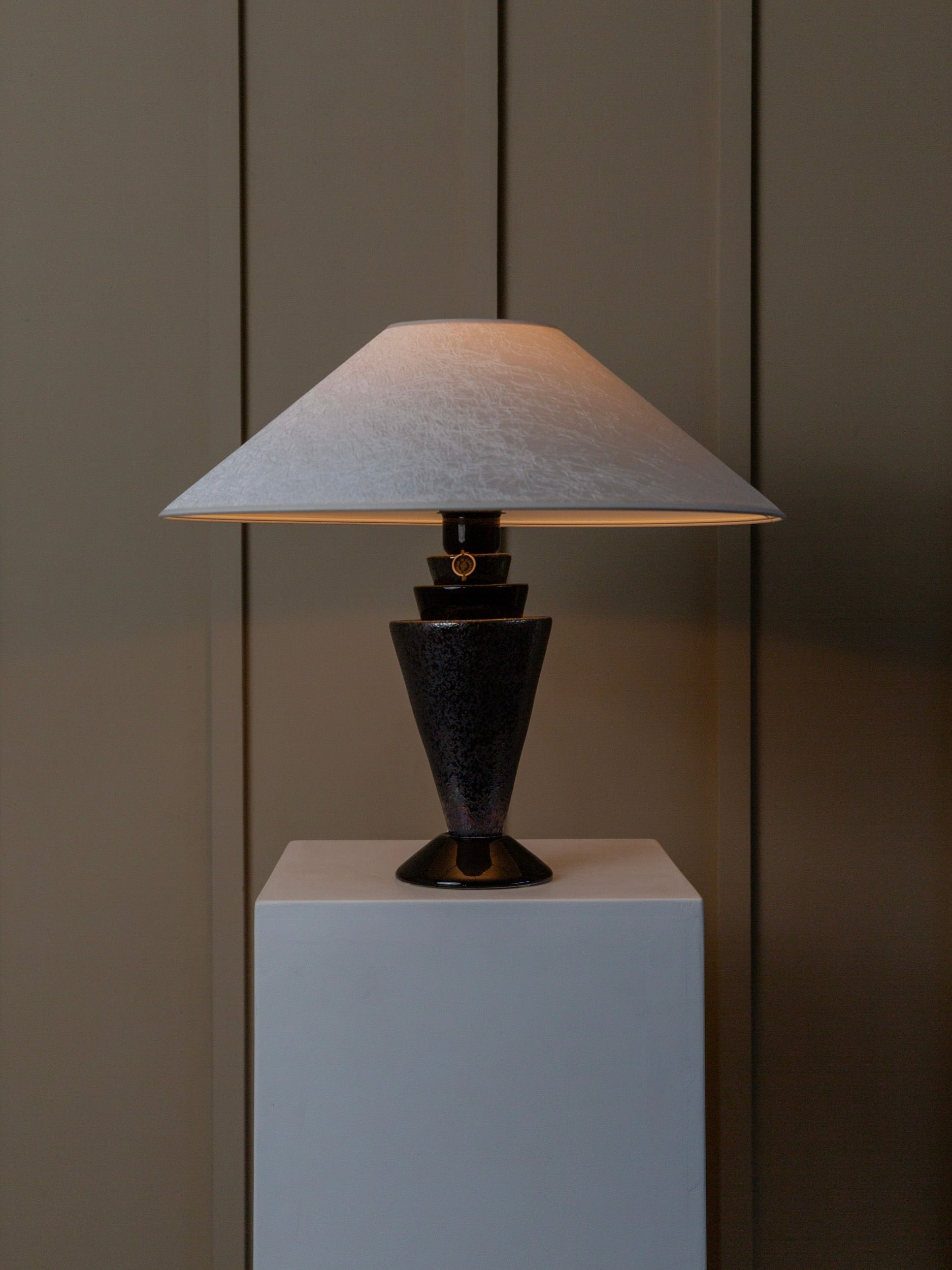 The Vintage Ceramic Table Lamp with Silk Shade - Italy 70s by Bottega Jacobs features a textured black base and a wide, conical white shade, adding stylish Hollywood Regency flair as it rests elegantly on a white pedestal against a muted, paneled wall.