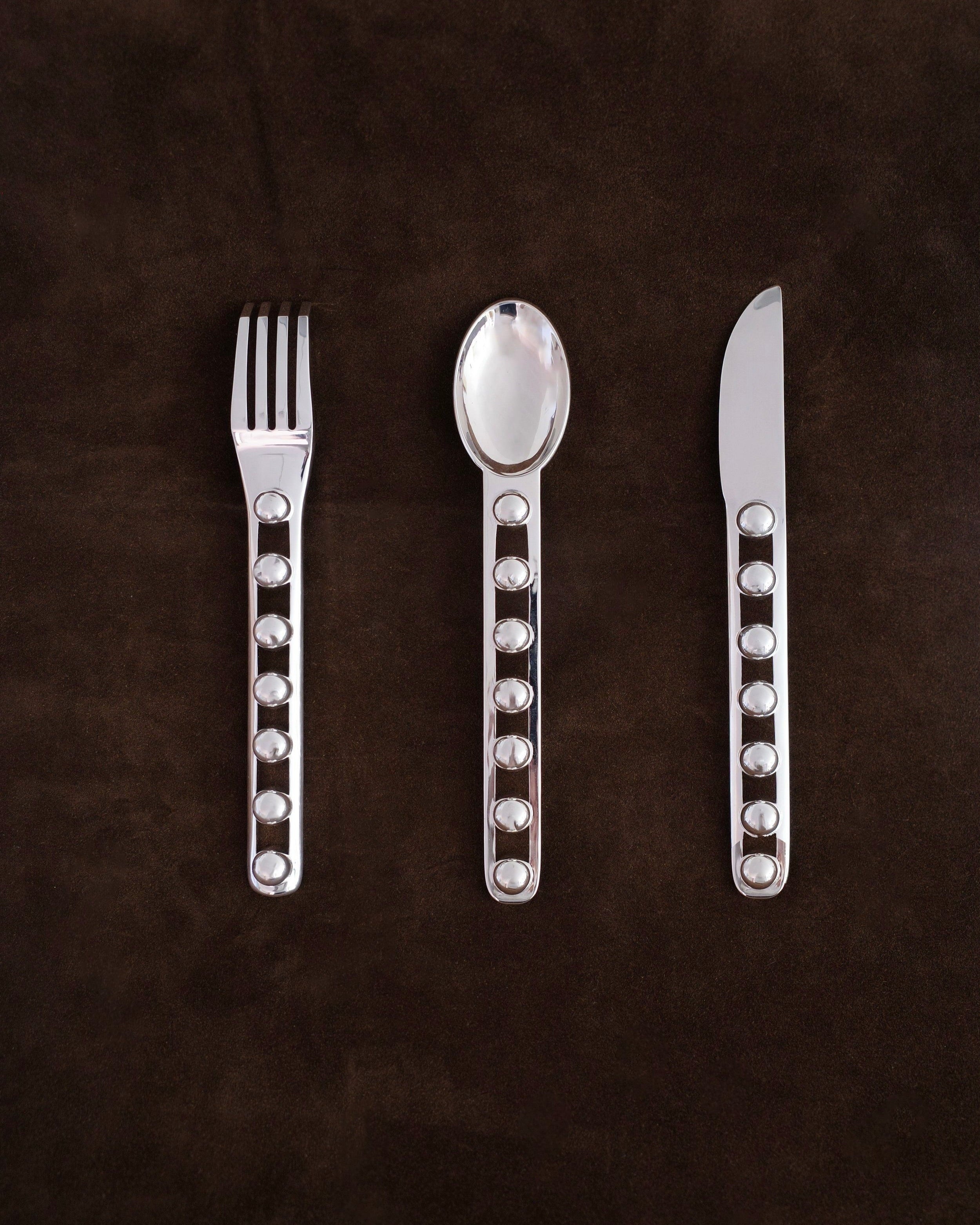 The "Seven Ball" Cutlery Set by Jessi Burch lies on a dark brown surface, showcasing a fork, spoon, and knife. The handles exhibit a design with circular cutouts, merging functionality with artistic elegance.