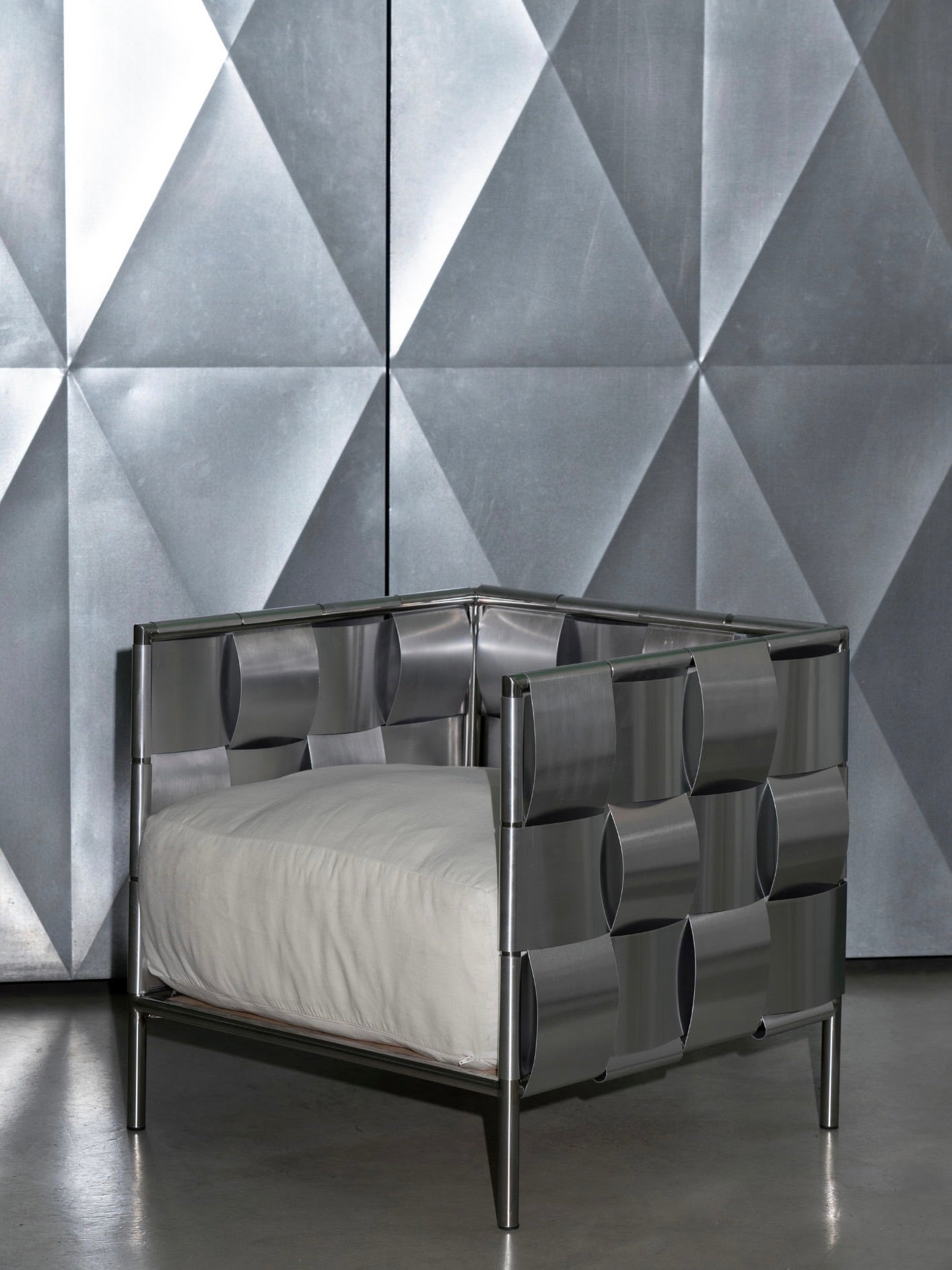 A modern stainless steel "Braid" Armchair from The Good Living&Co. with a geometric design featuring oval cutouts stands on a concrete floor, showcasing a white cushion against a metallic, diamond-patterned backdrop.